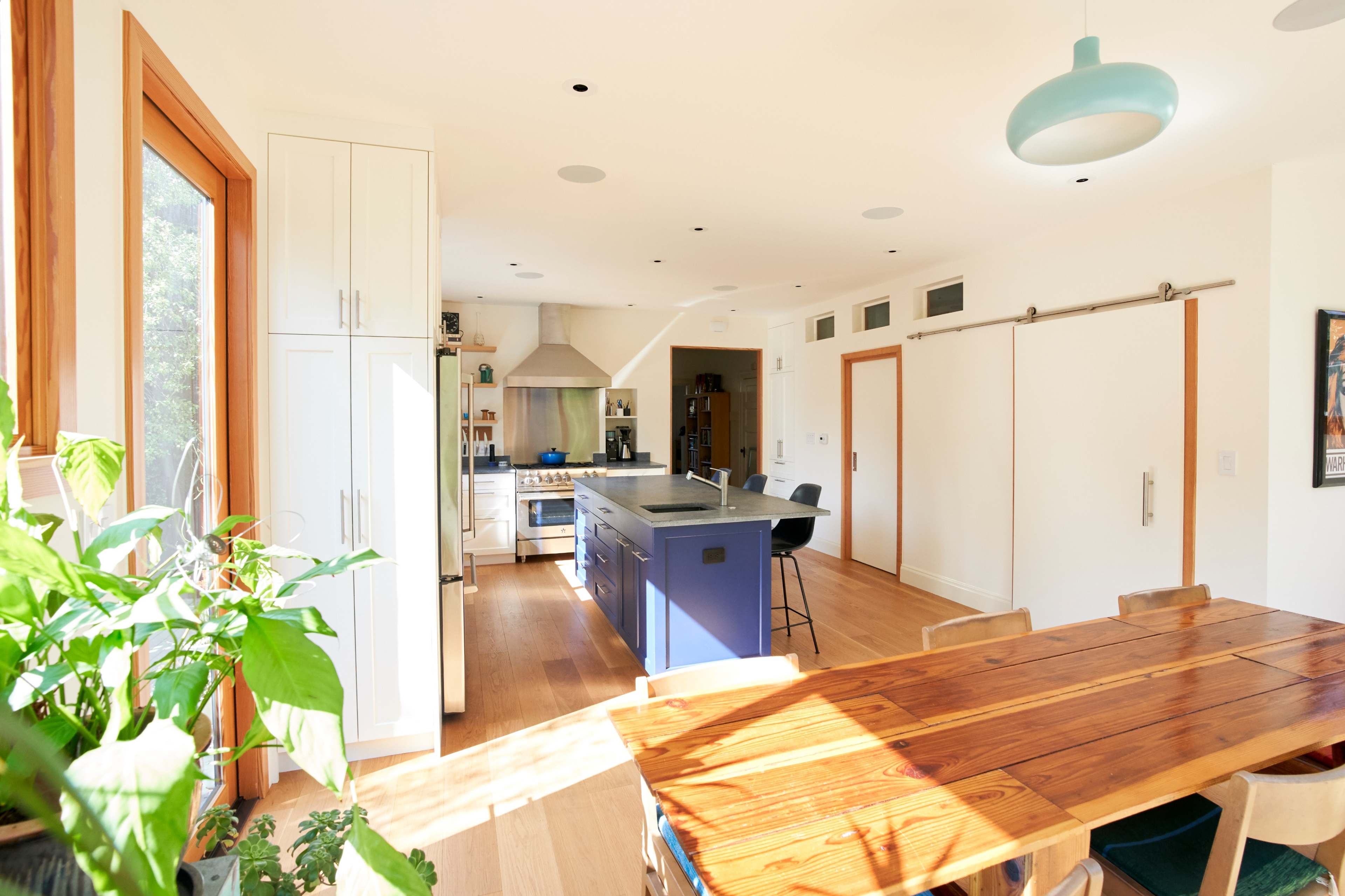 A modern kitchen features a central island with a blue base, white cabinetry, a wooden dining table, and large windows that allow natural light to enter.