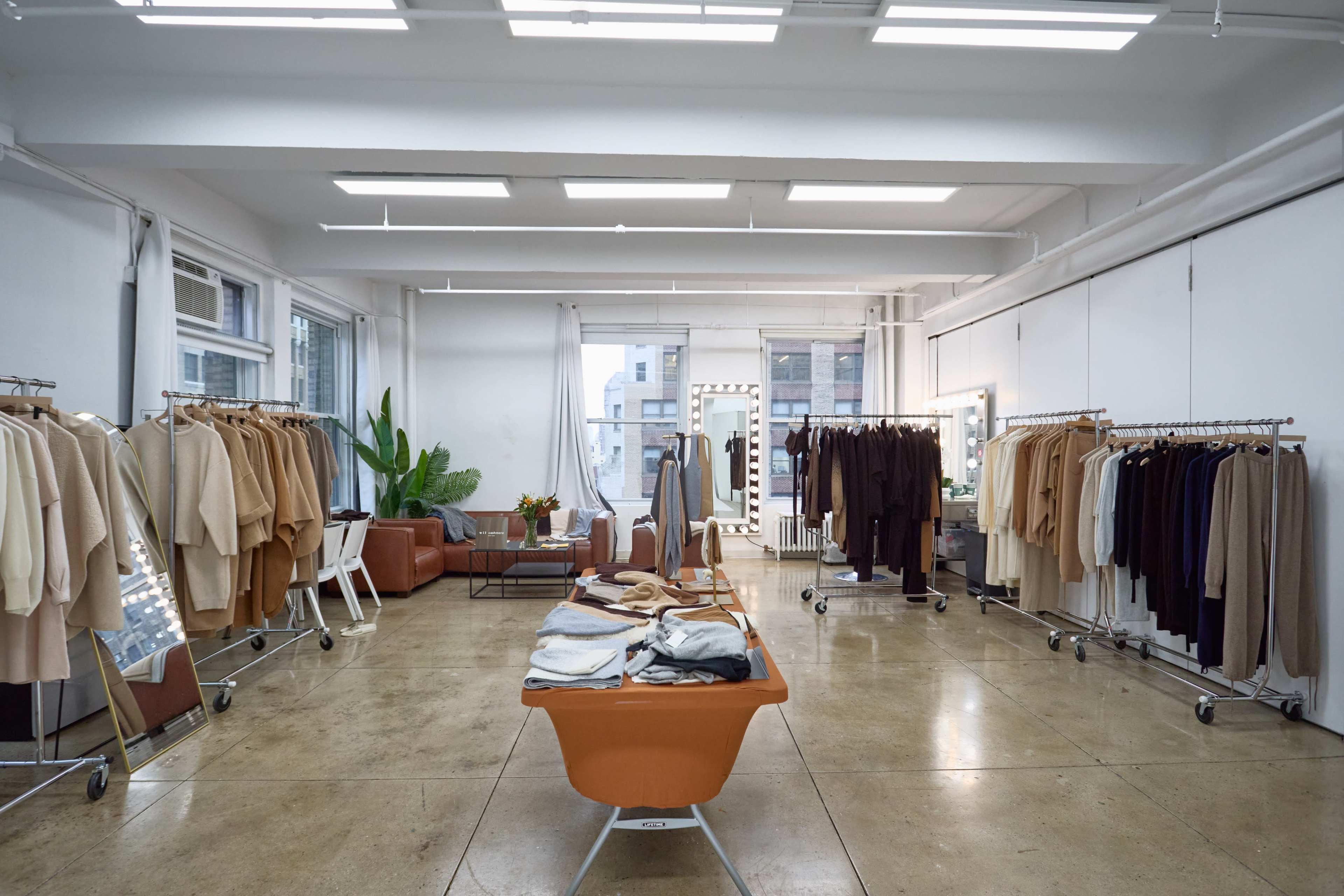 The image shows a minimalist clothing showroom featuring clothes on racks, a table with folded garments, a seating area, and large windows allowing natural light.