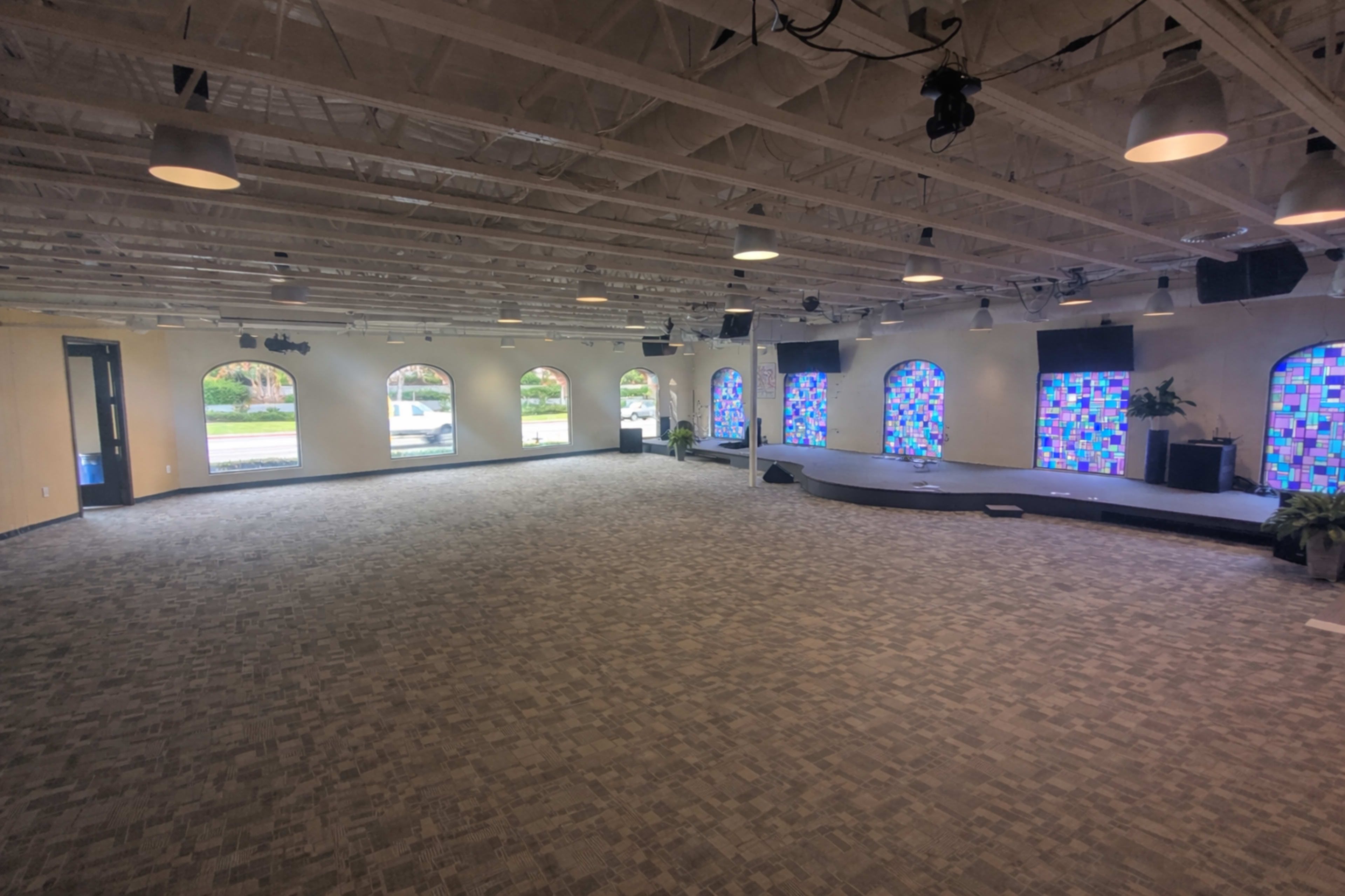 The image shows a spacious, well-lit interior room with a carpeted floor, featuring several arched windows with colorful stained glass and a small raised platform at one end.