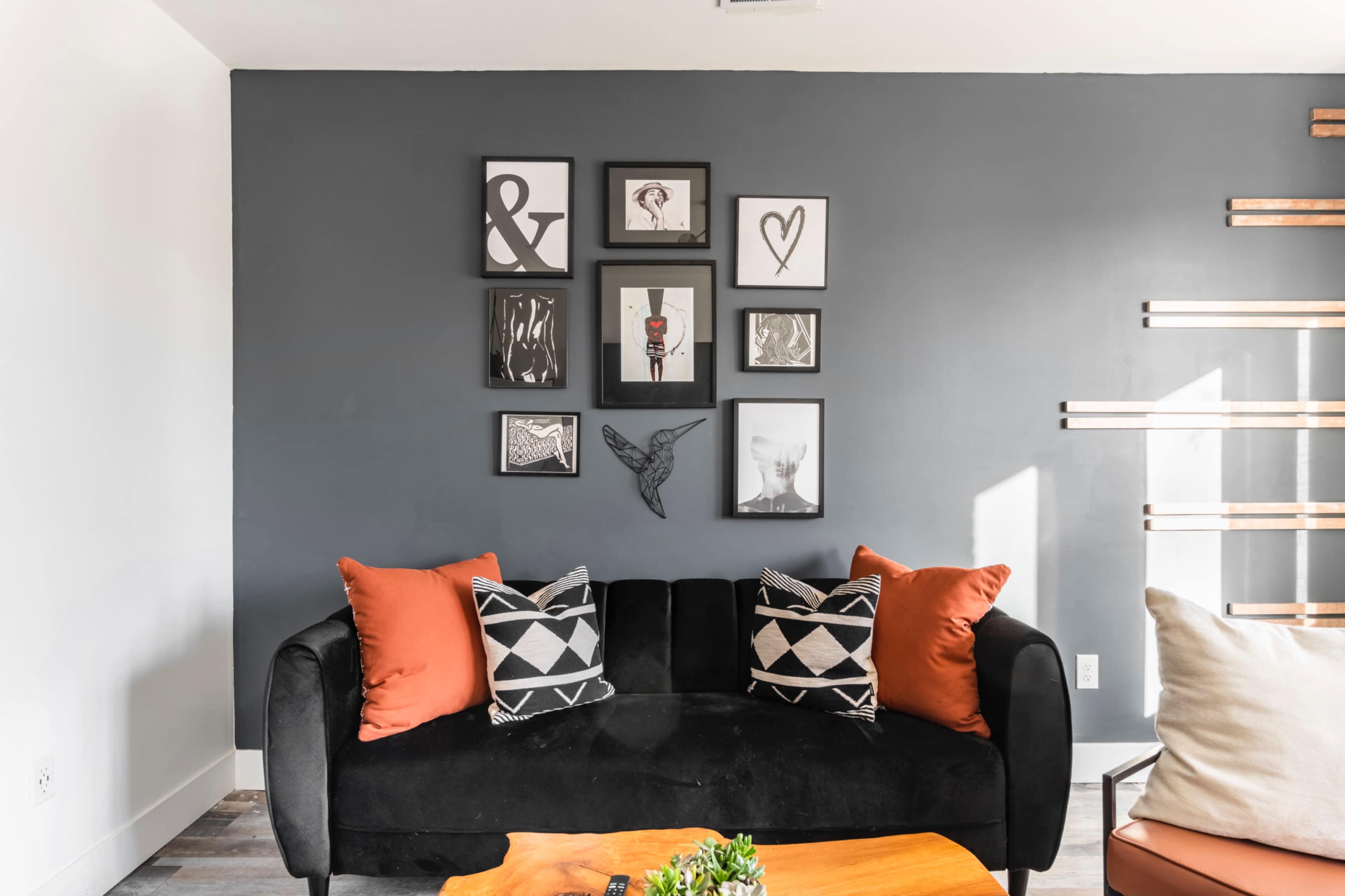 A black sofa with decorative pillows in front of a dark gray wall adorned with a series of framed artworks and a wooden display unit.