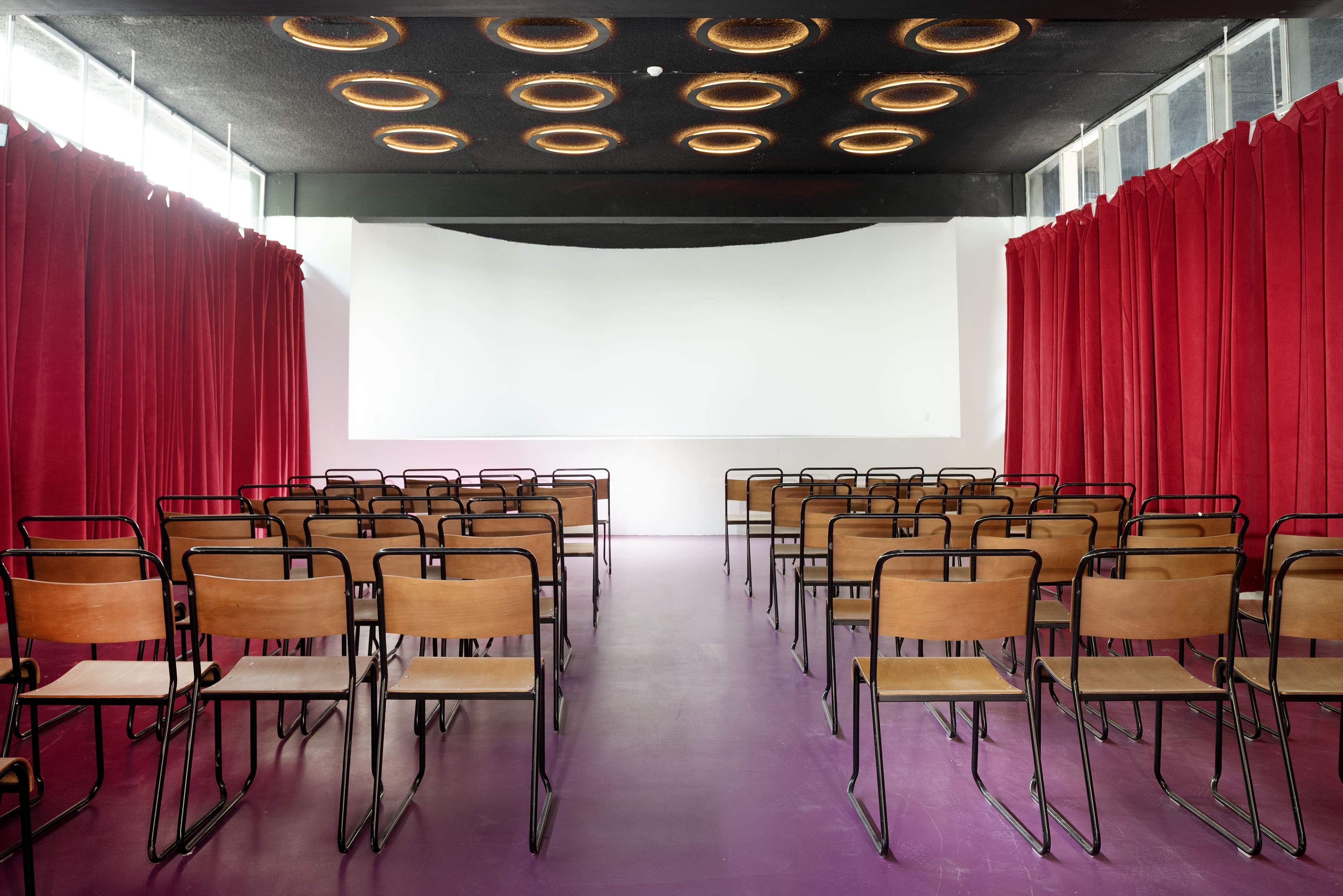 The image shows a spacious seminar room with rows of metal-framed wooden chairs facing a blank white screen, flanked by red curtains.