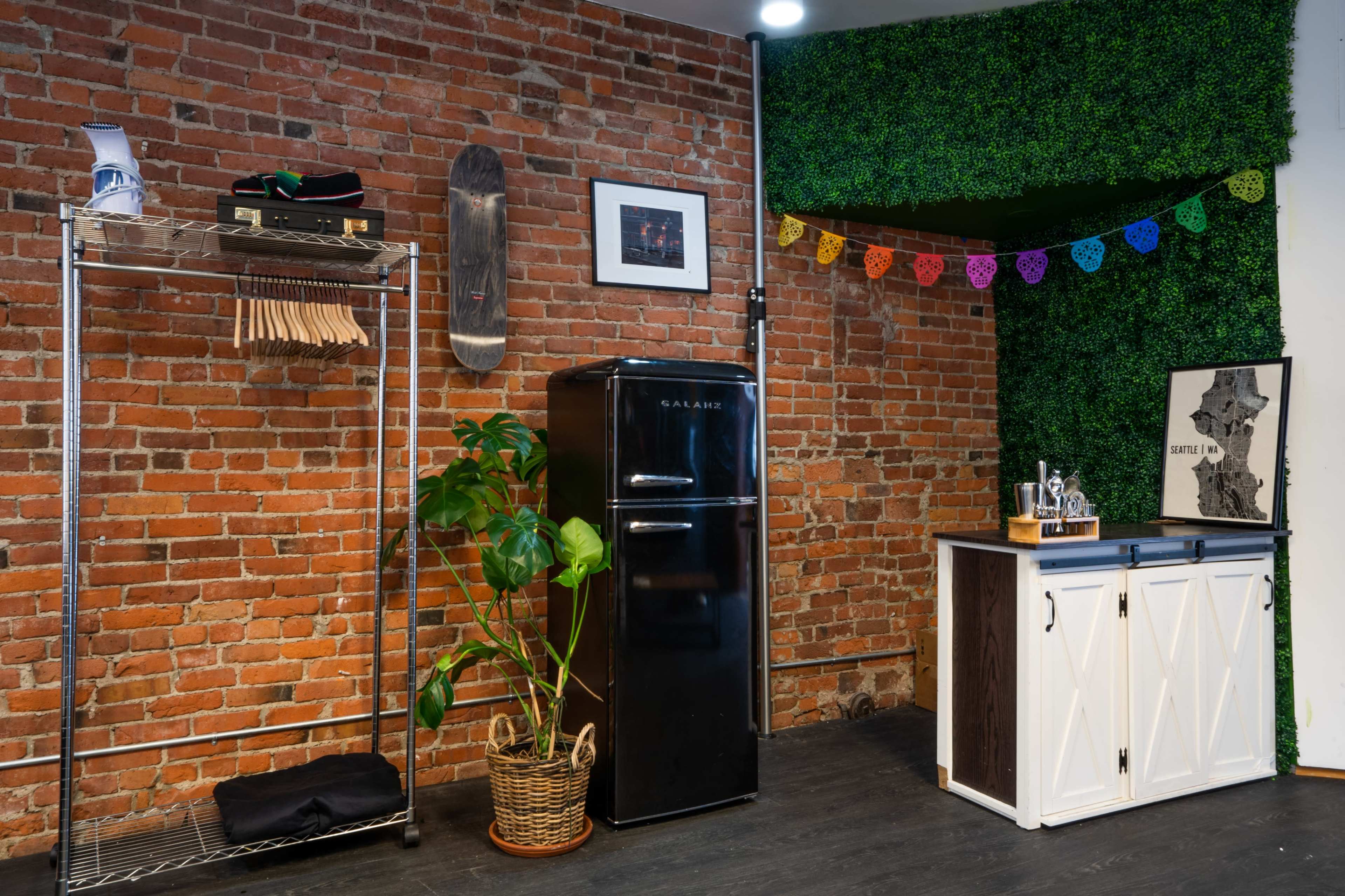 A room with a brick wall, featuring a black refrigerator, a metal rack with wooden hangers, a potted plant, and a wooden cabinet adorned with decorative banners.