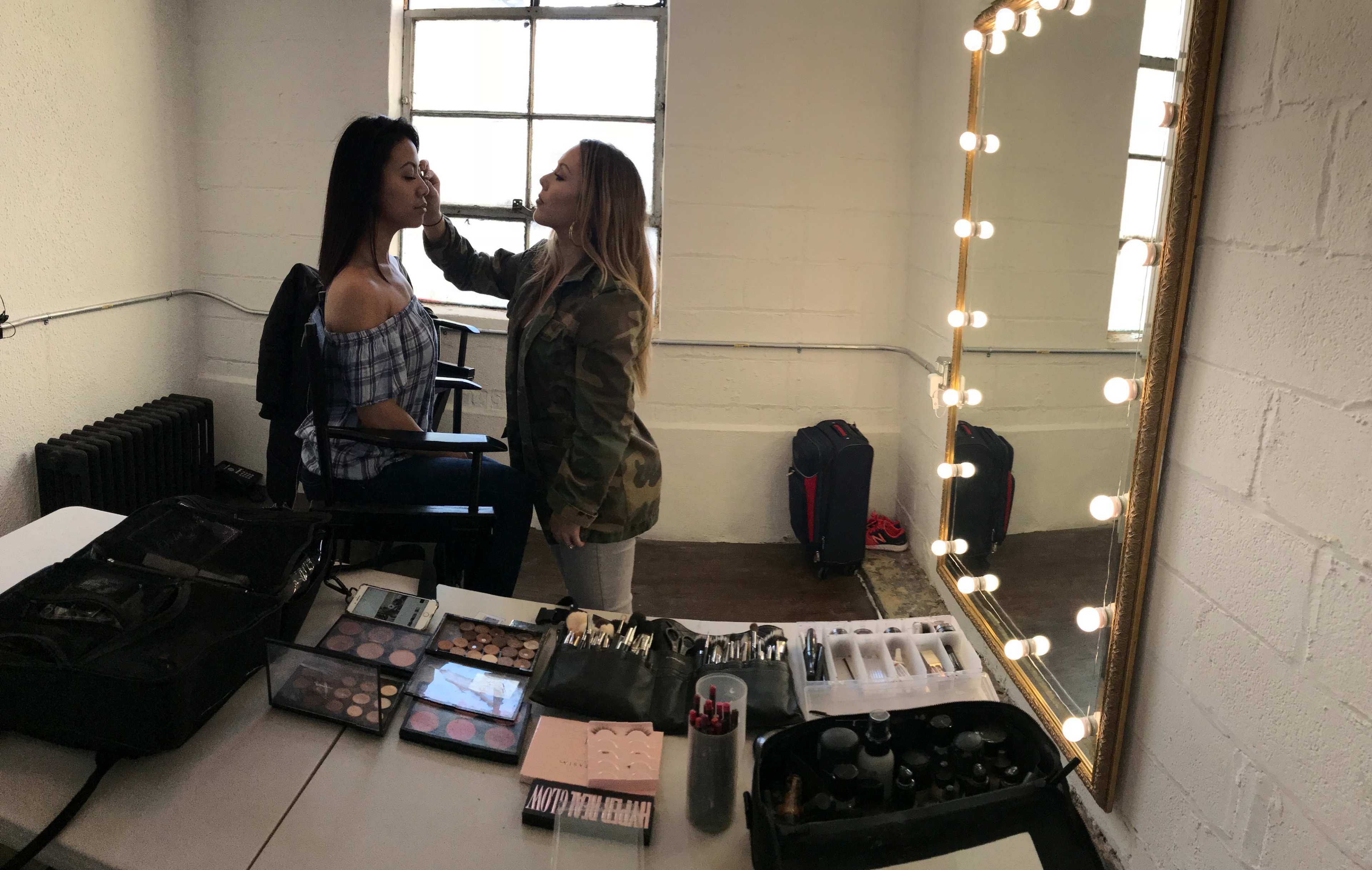 A makeup artist applies cosmetics to a model in a well-lit studio with a large mirror and multiple makeup products displayed on a table.