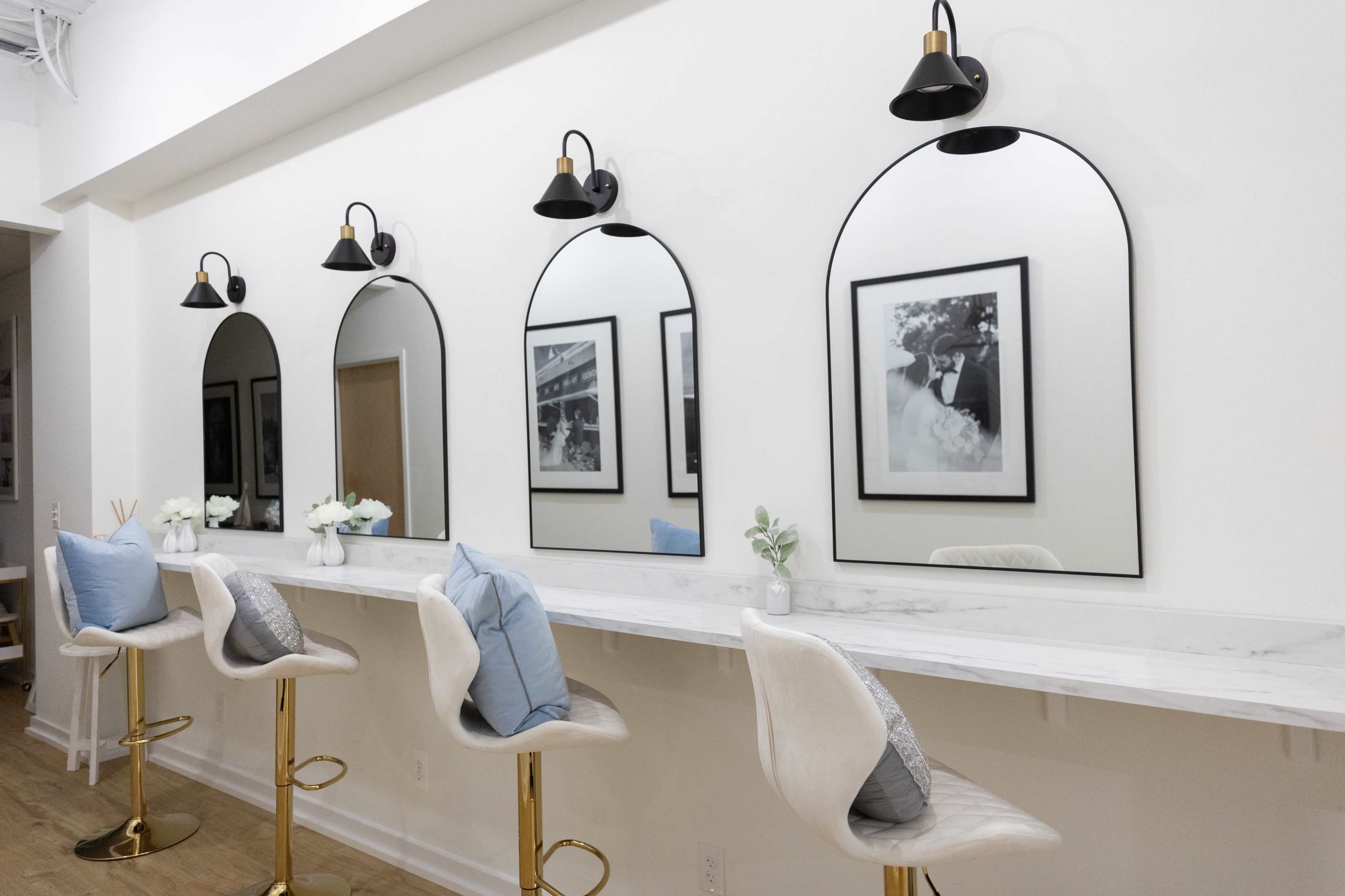 The image features a modern vanity area with four arched mirrors, stylish black sconces, and minimalist bar stools arranged along a marble counter.