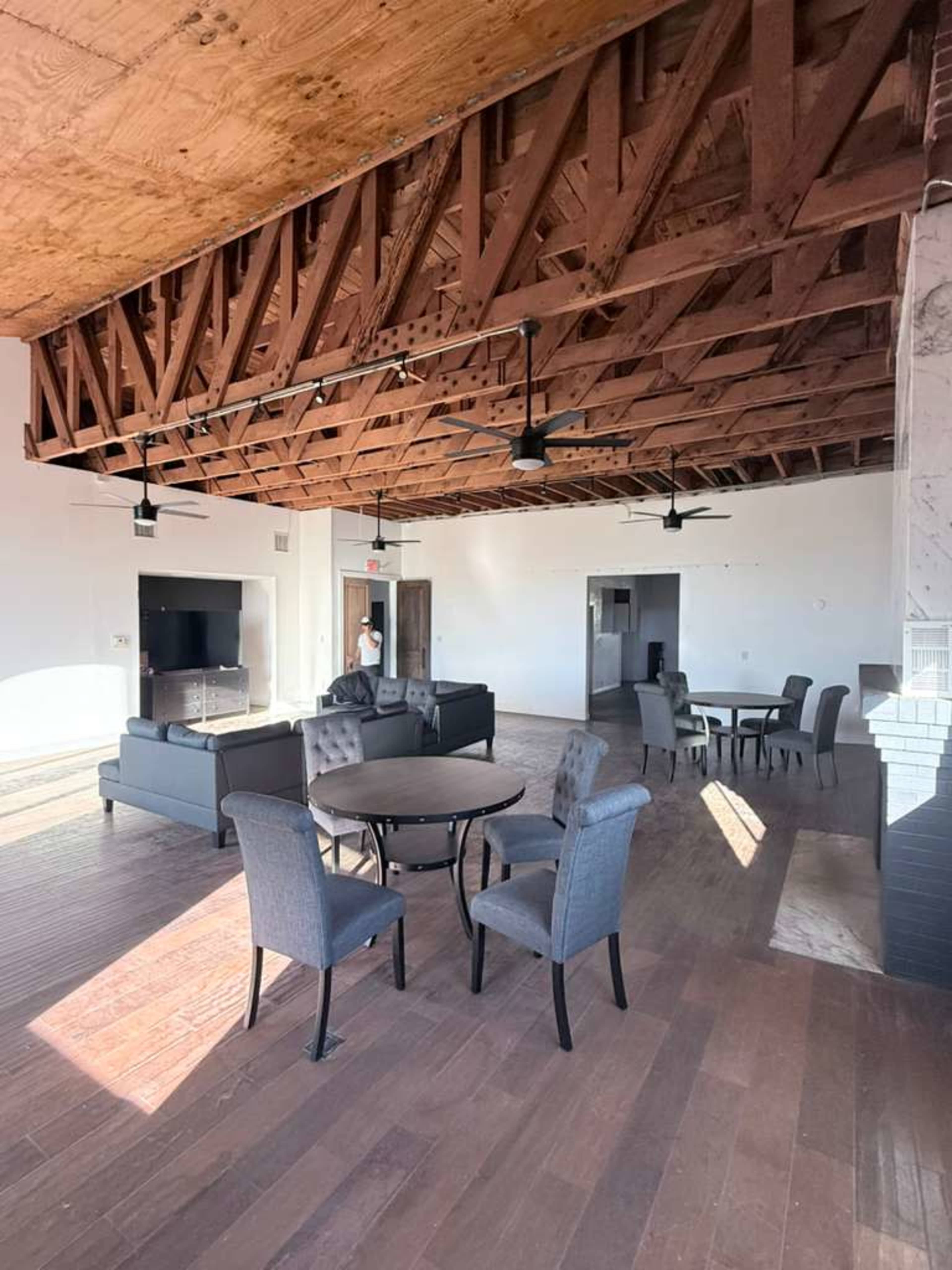 The image shows a spacious open area with a high wooden ceiling, featuring a seating arrangement that includes a sofa, armchairs, and a dining table surrounded by chairs.