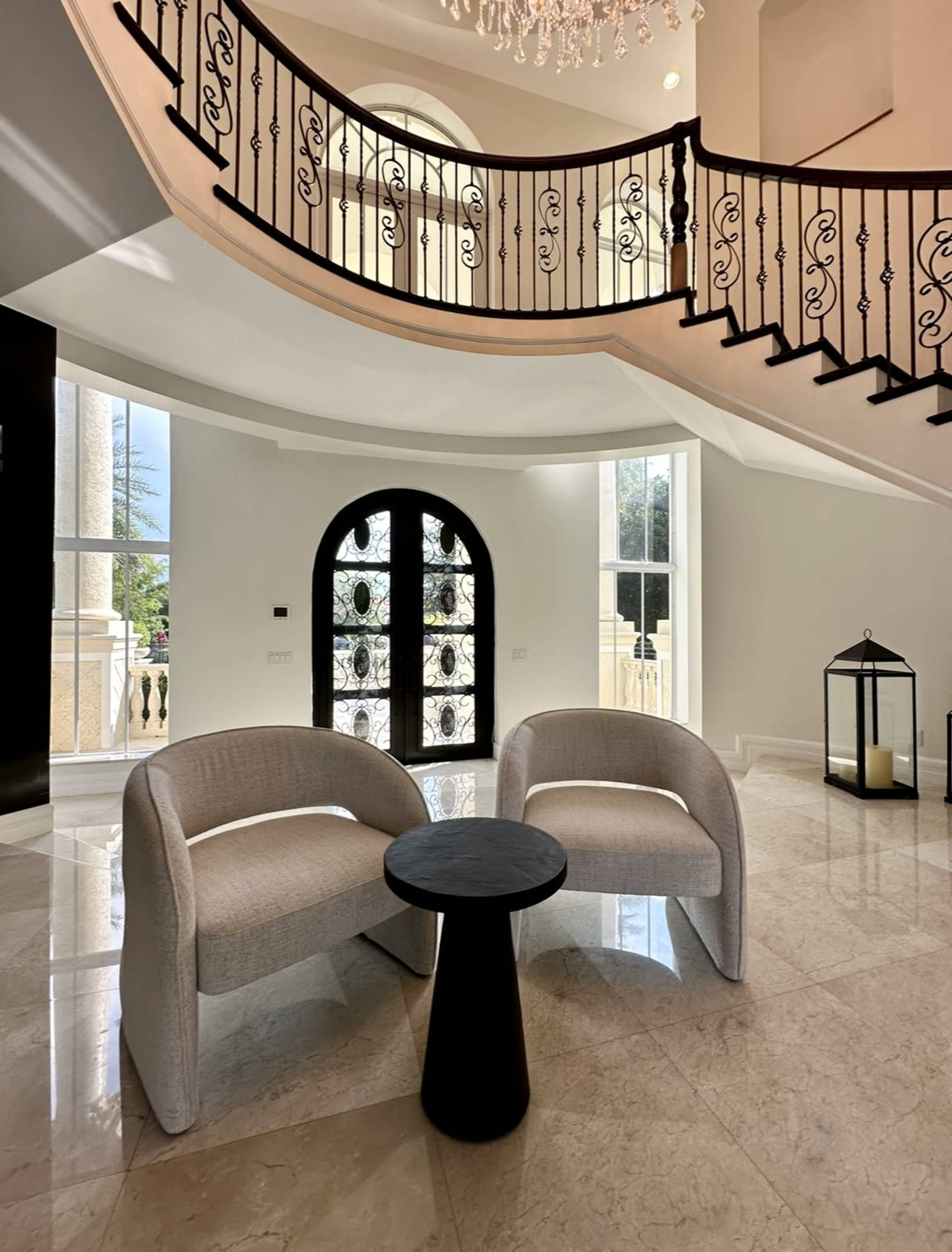 Two upholstered chairs with a black side table, set in a spacious foyer that includes a staircase and large windows.