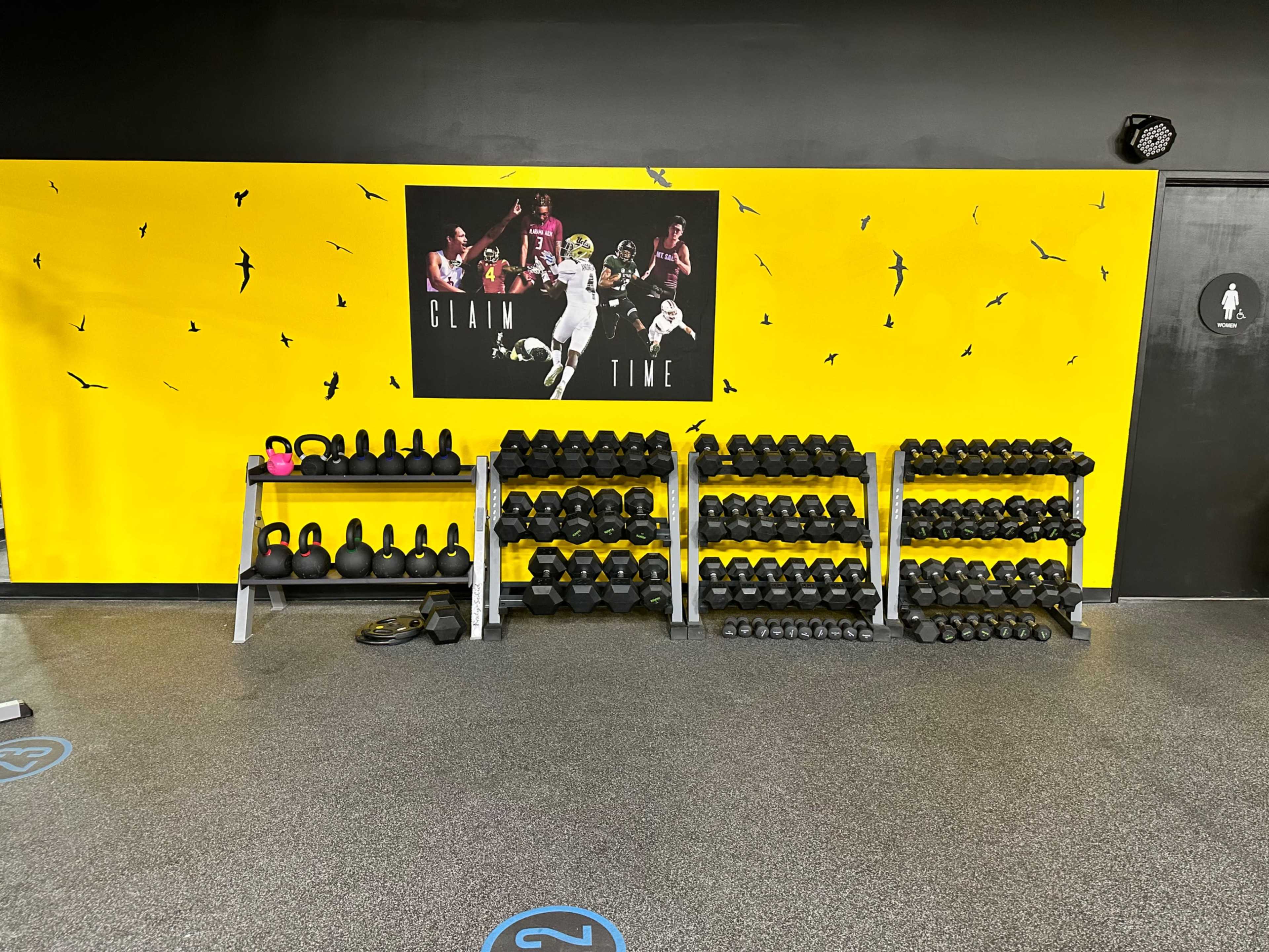 A fitness area featuring multiple racks of dumbbells and kettlebells against a vibrant yellow wall with a motivational poster.