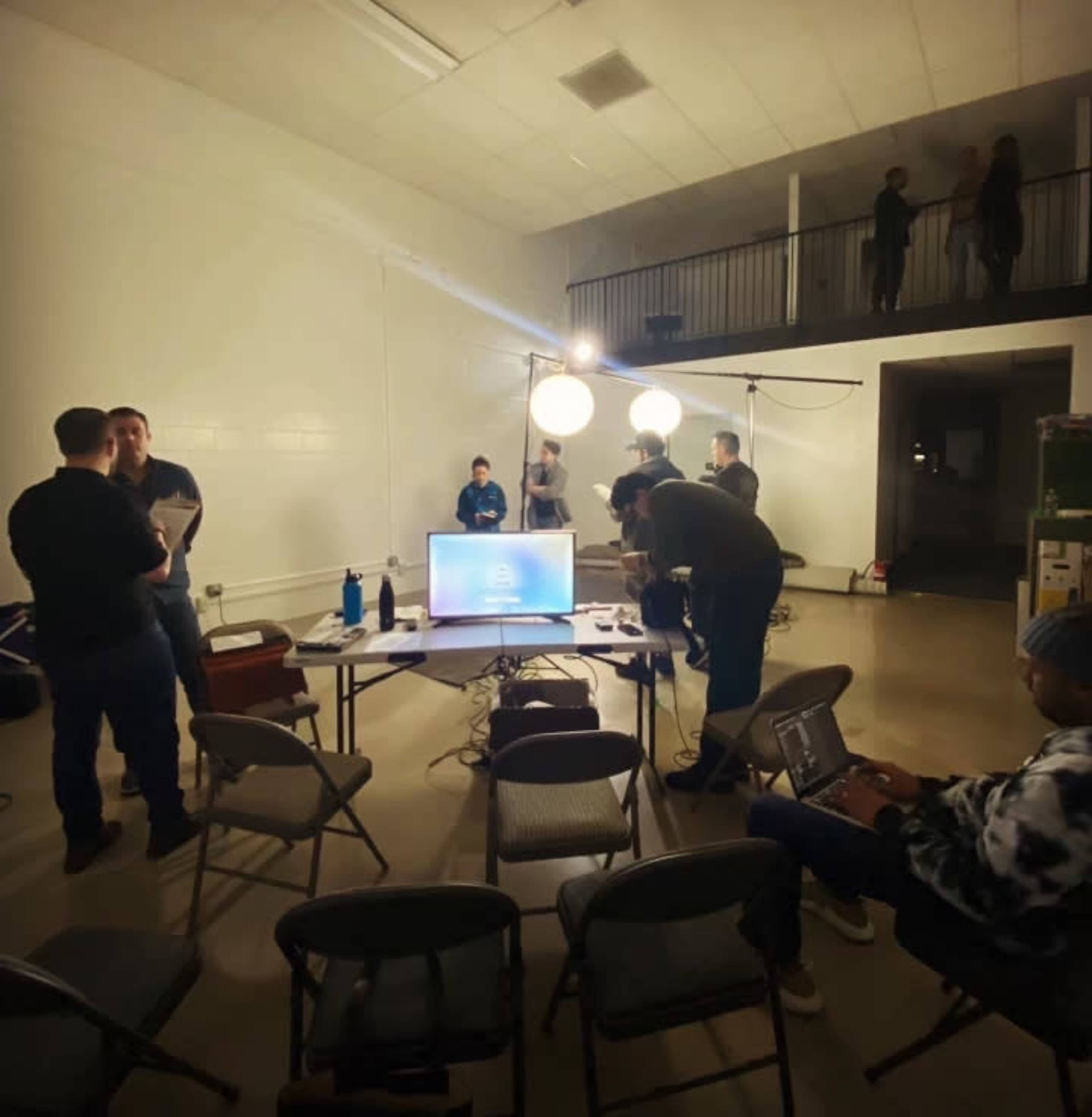 A group of people is gathered in a well-lit room, working around a table with a computer while others adjust equipment and set up lighting.