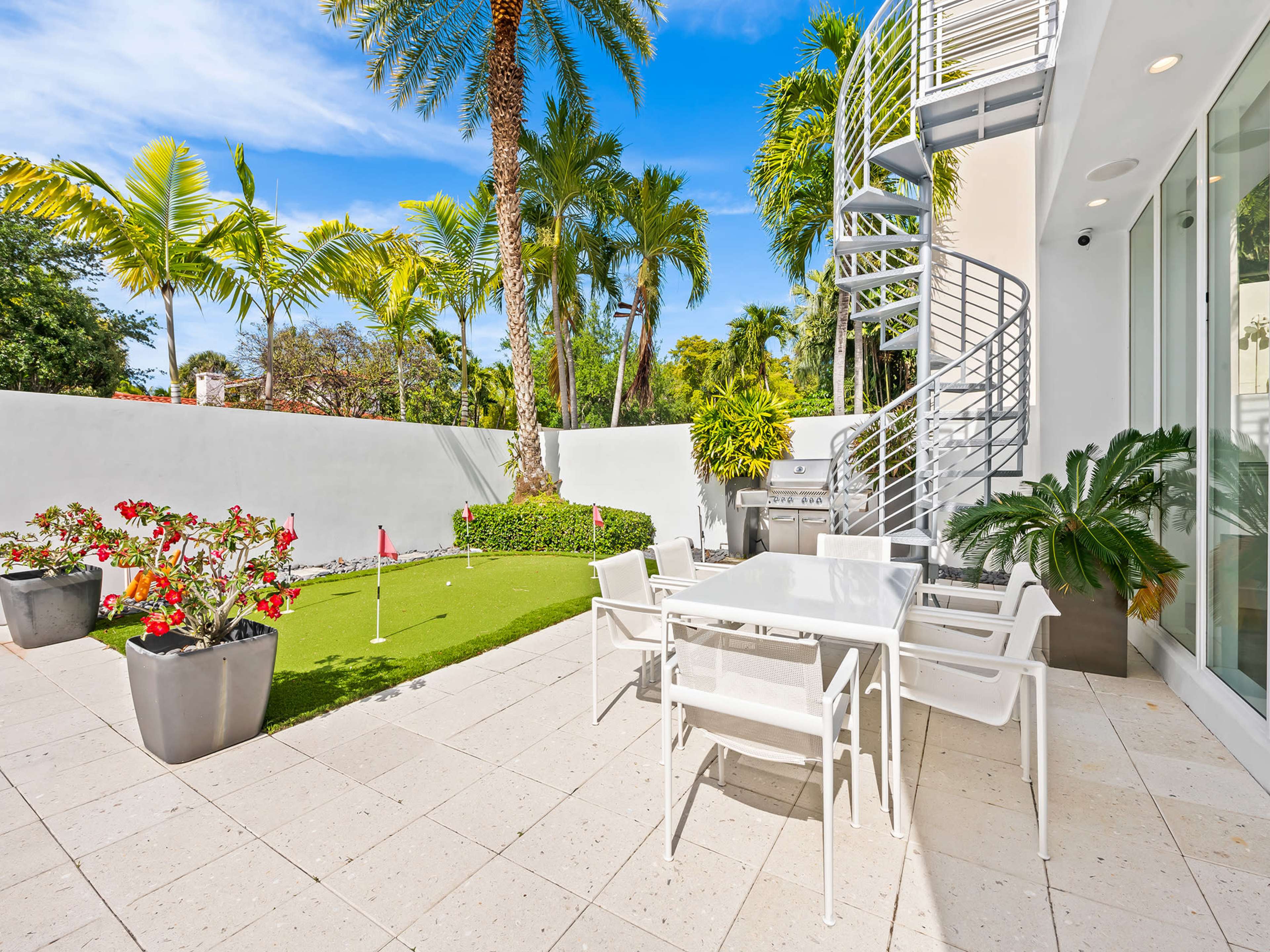 The image shows a well-lit outdoor patio area with a spiral staircase, a small putting green, potted plants, and a dining table set for six.