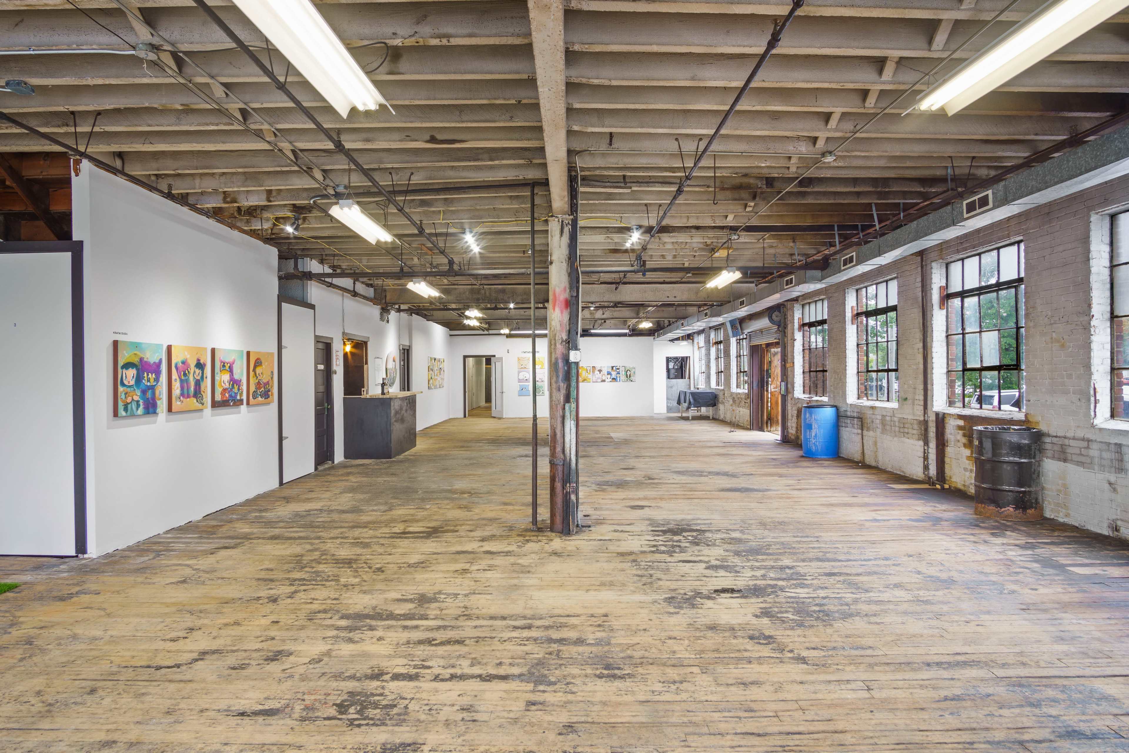 An empty art gallery space with white walls, wooden floors, and large windows, illuminated by overhead lights.