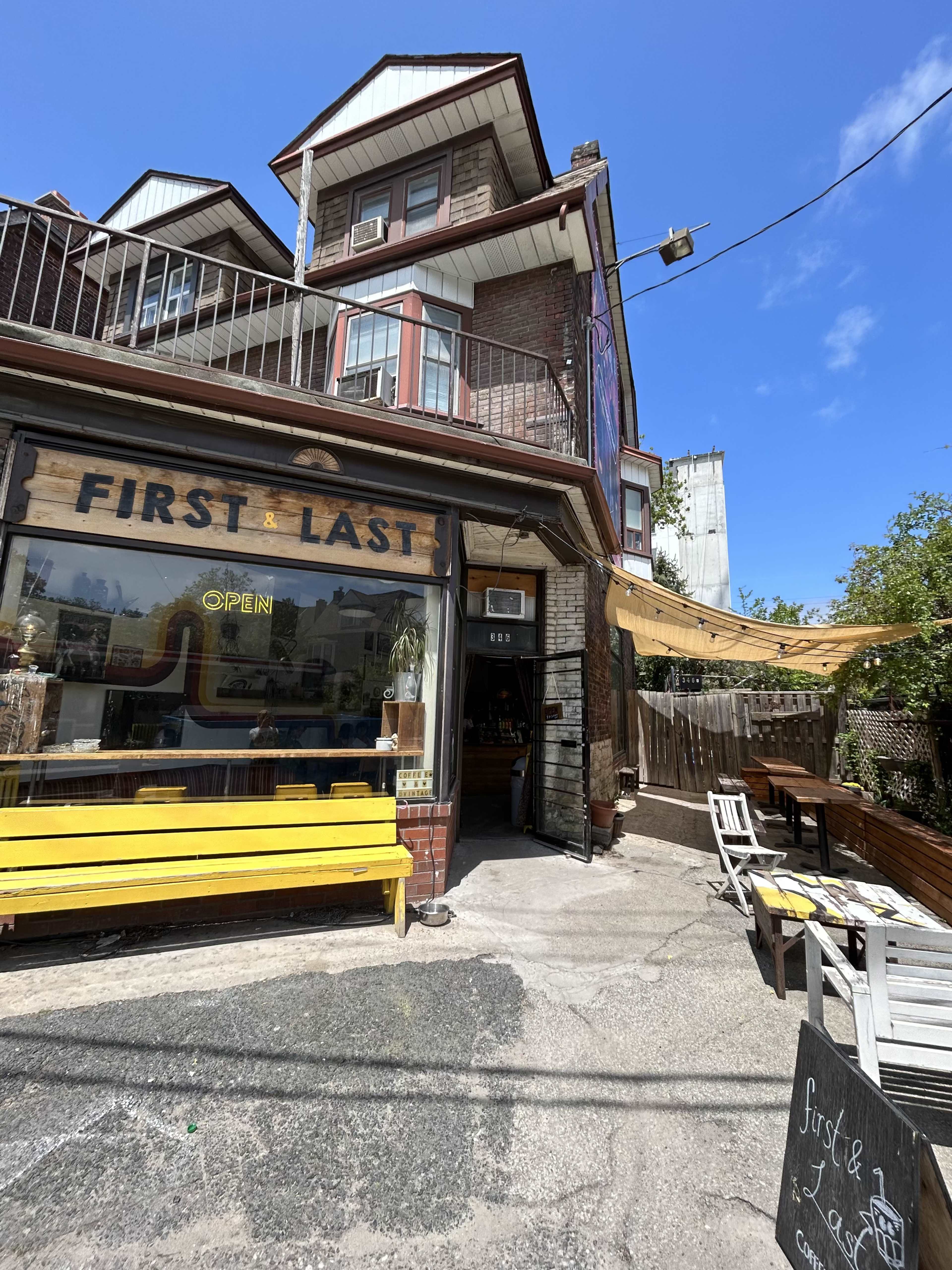 A café named "First & Last" with a yellow bench outside, situated in a brick building with multiple stories and an open sign.