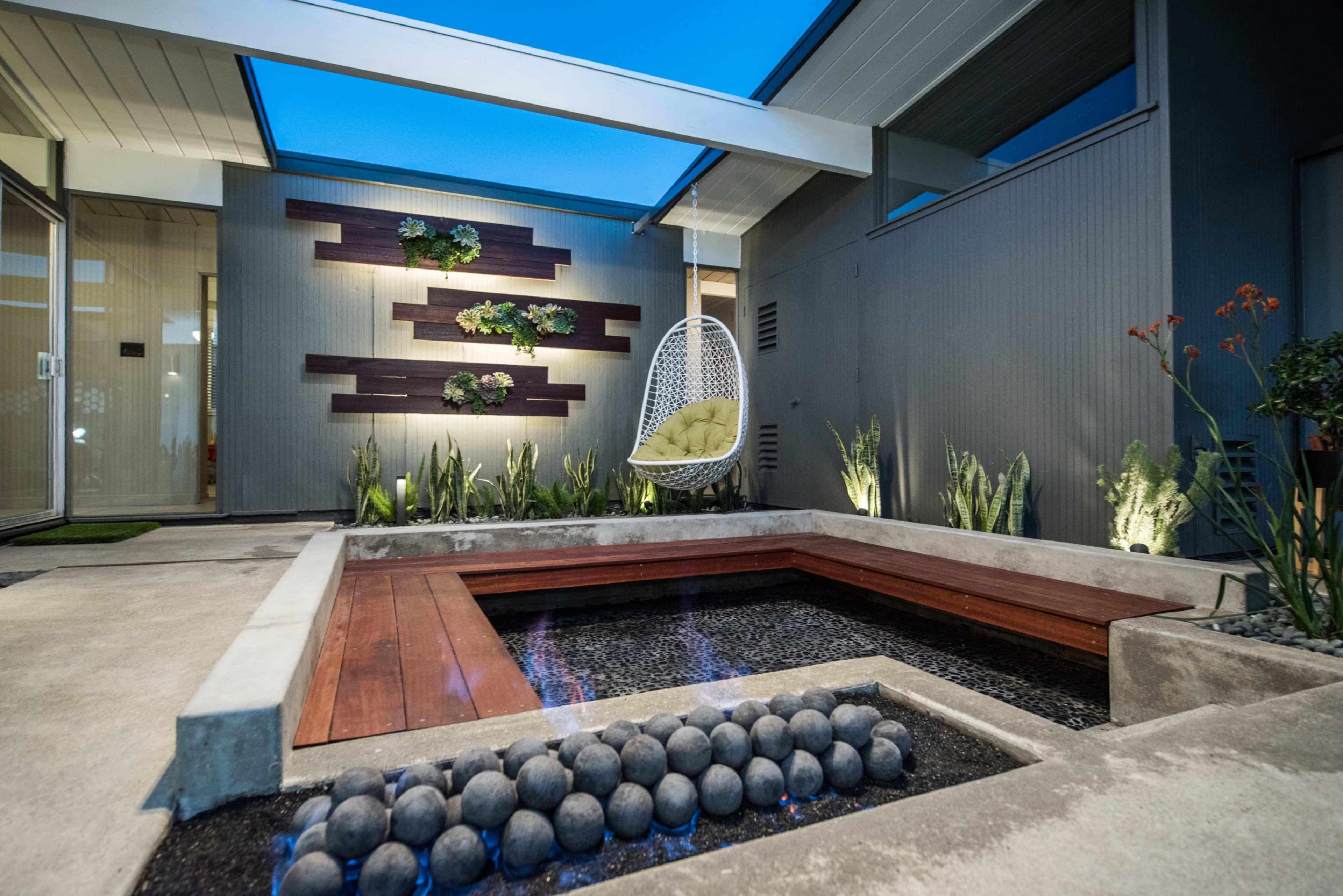 A modern outdoor space featuring a hanging chair, a small water feature with round stones, and vertical garden elements against a sleek wall.
