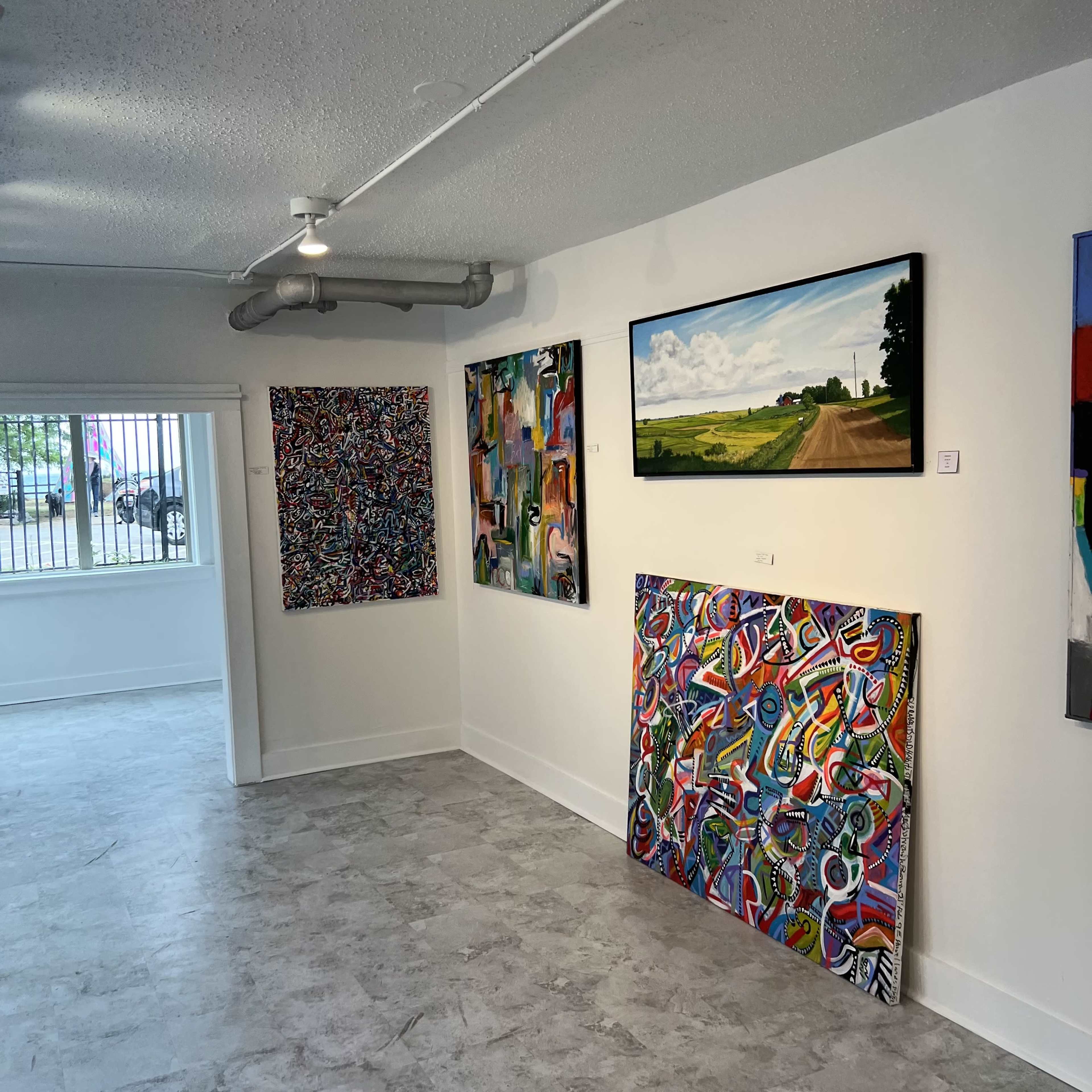 The image shows an art gallery with colorful paintings displayed on white walls and a light gray floor.