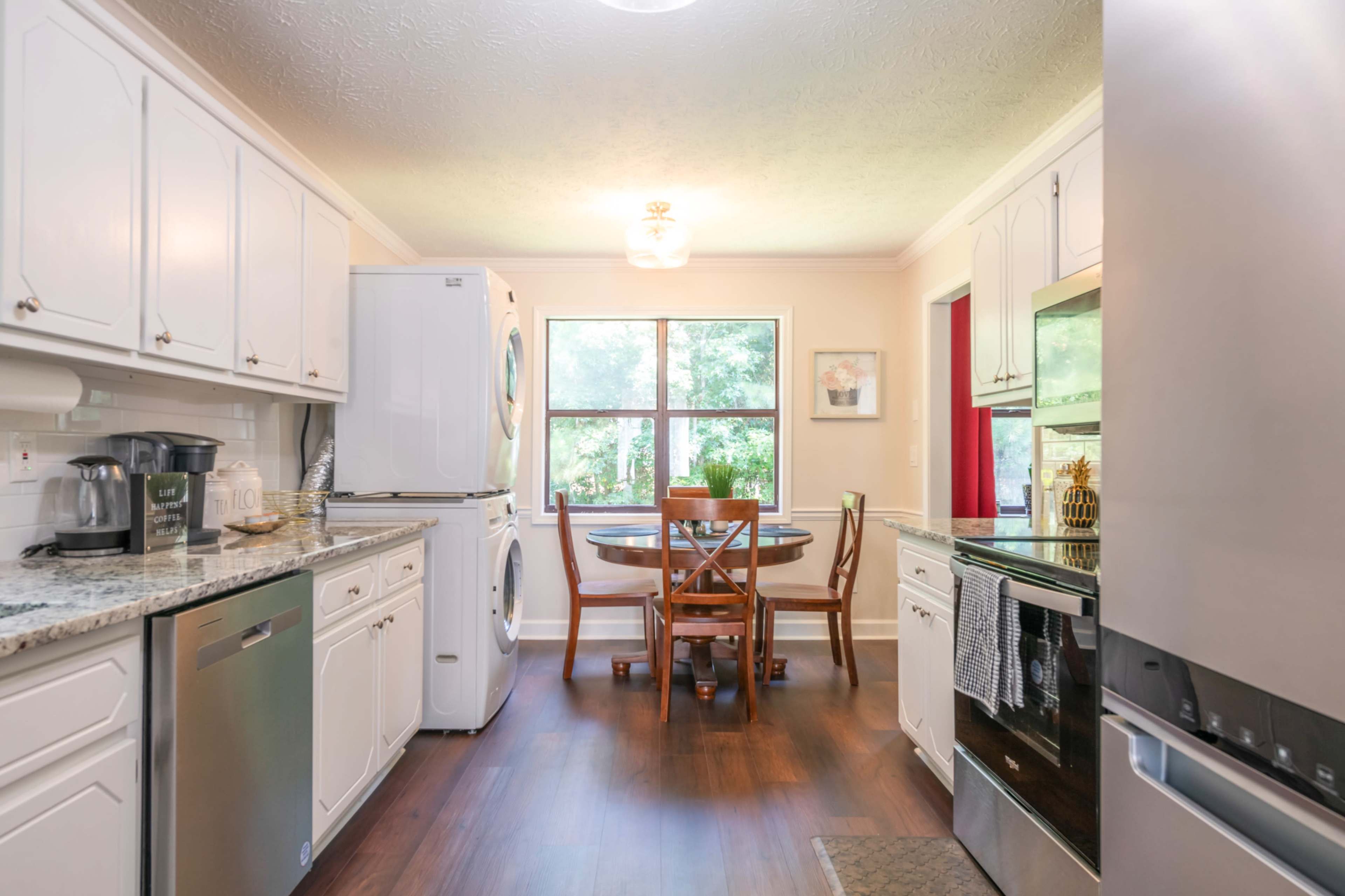 A bright kitchen features white cabinets, a round wooden table with four chairs, and a washer and dryer near a window overlooking greenery.