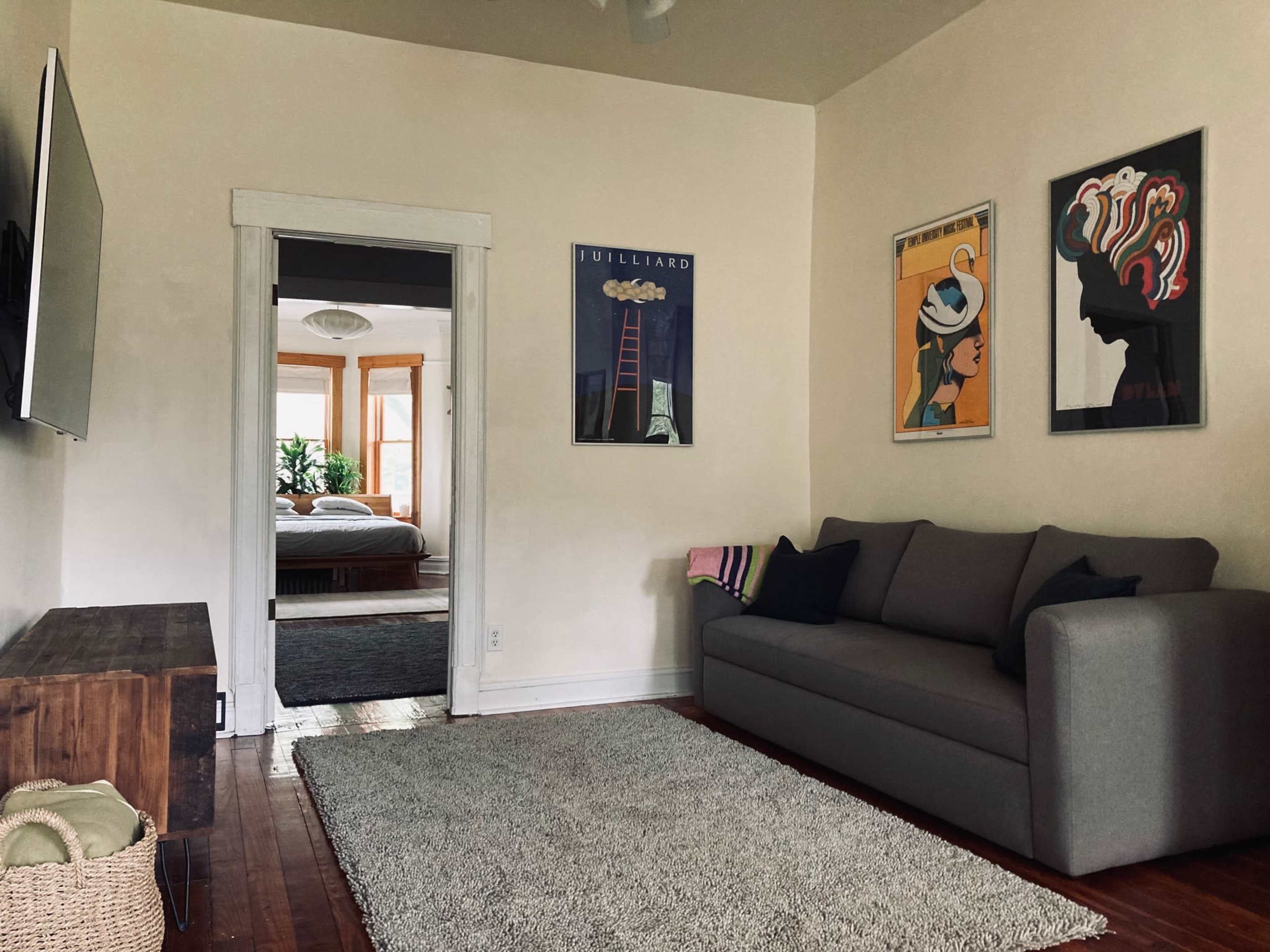 The image shows a living room with a gray sofa, a large area rug, and wall art, leading to a doorway that opens into a bedroom.