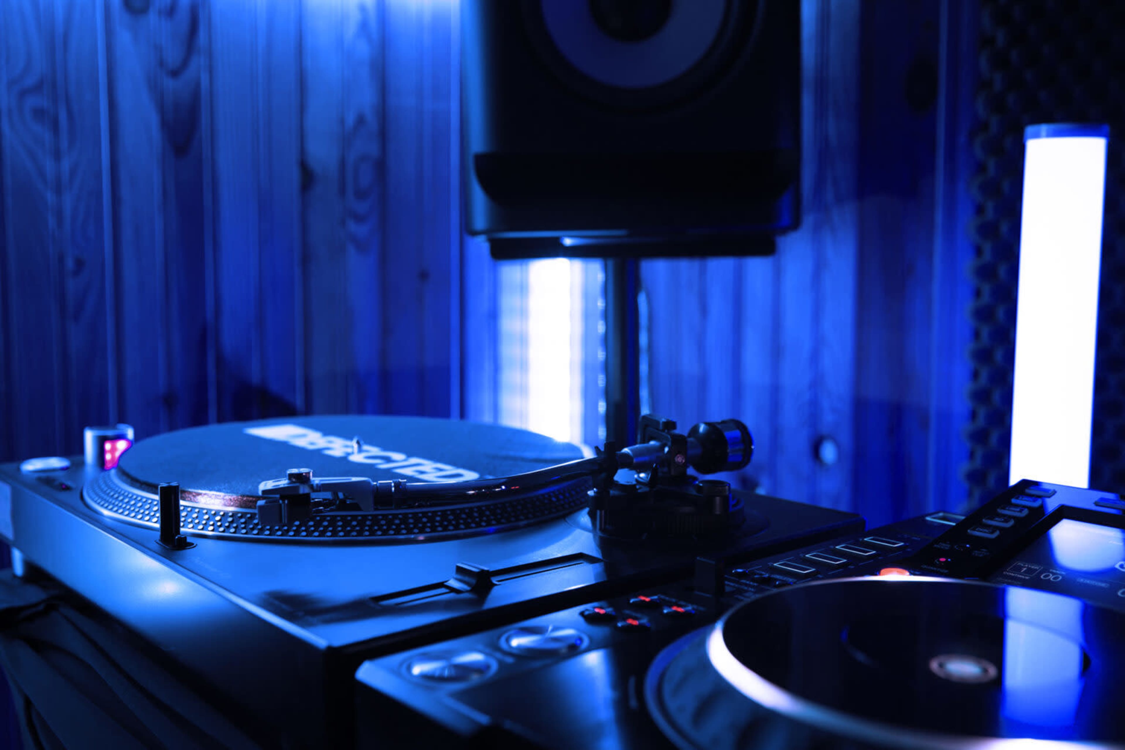 A close-up of a DJ setup featuring turntables and a mixer, illuminated by blue lighting in a wooden-paneled room.