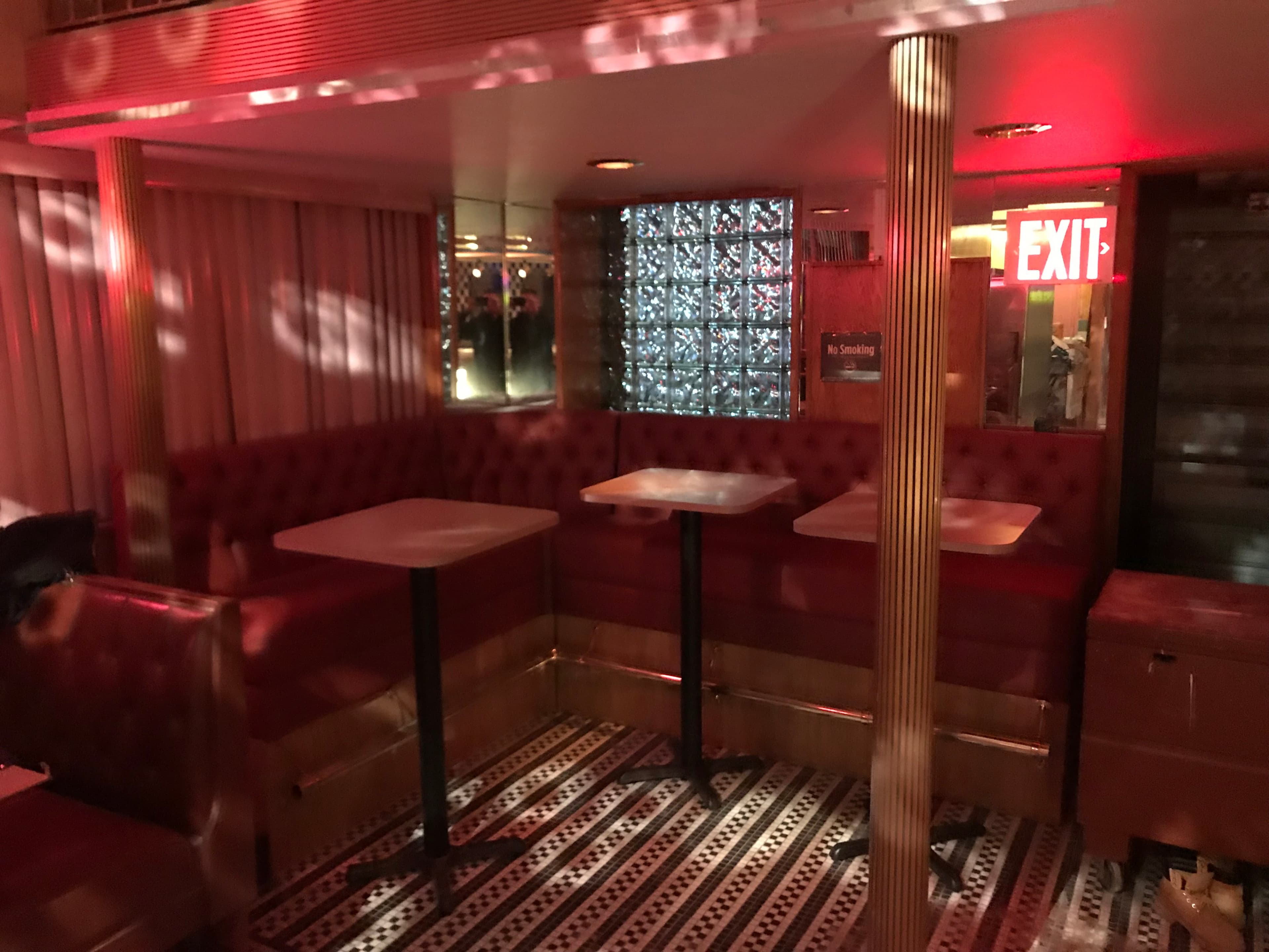 A dimly lit bar area with red lighting, featuring red tufted seating, small tables, and a glass block wall near an exit sign.