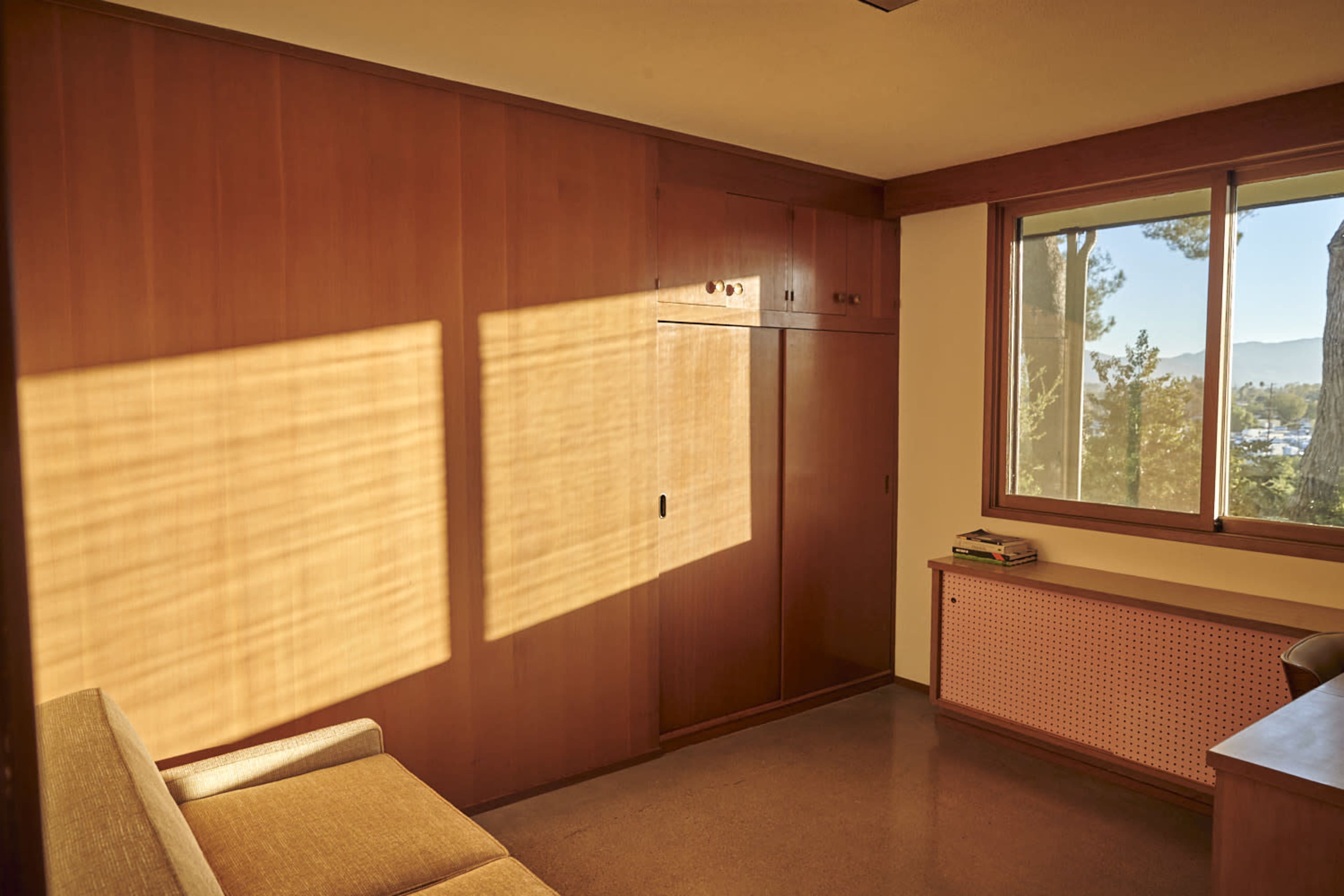 A room features wooden walls, a window with sunlight casting shadows, and minimal furnishings including a sofa and a cabinet.