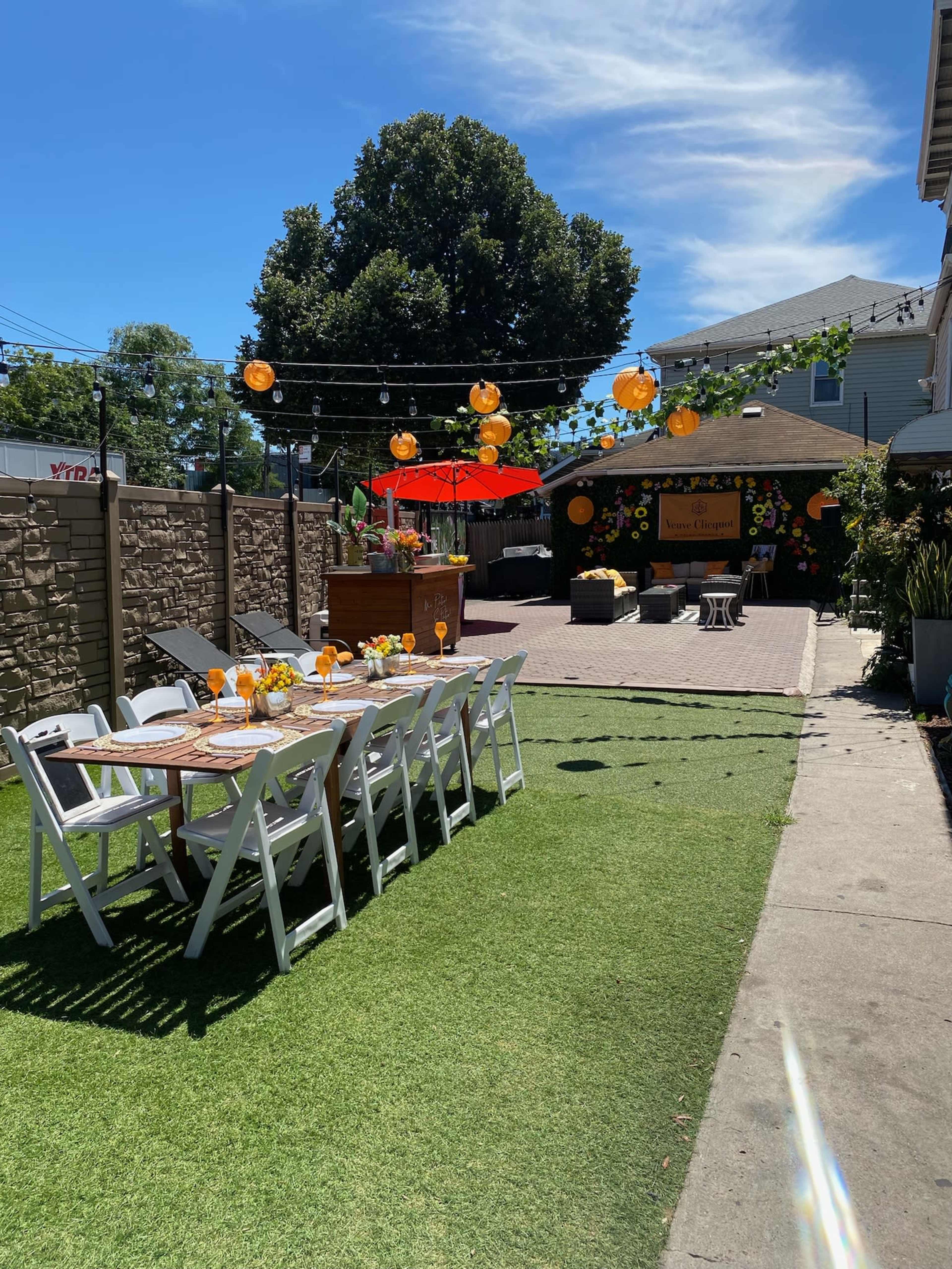 A decorated outdoor space features a long dining table set with flowers, surrounded by lush greenery and string lights.