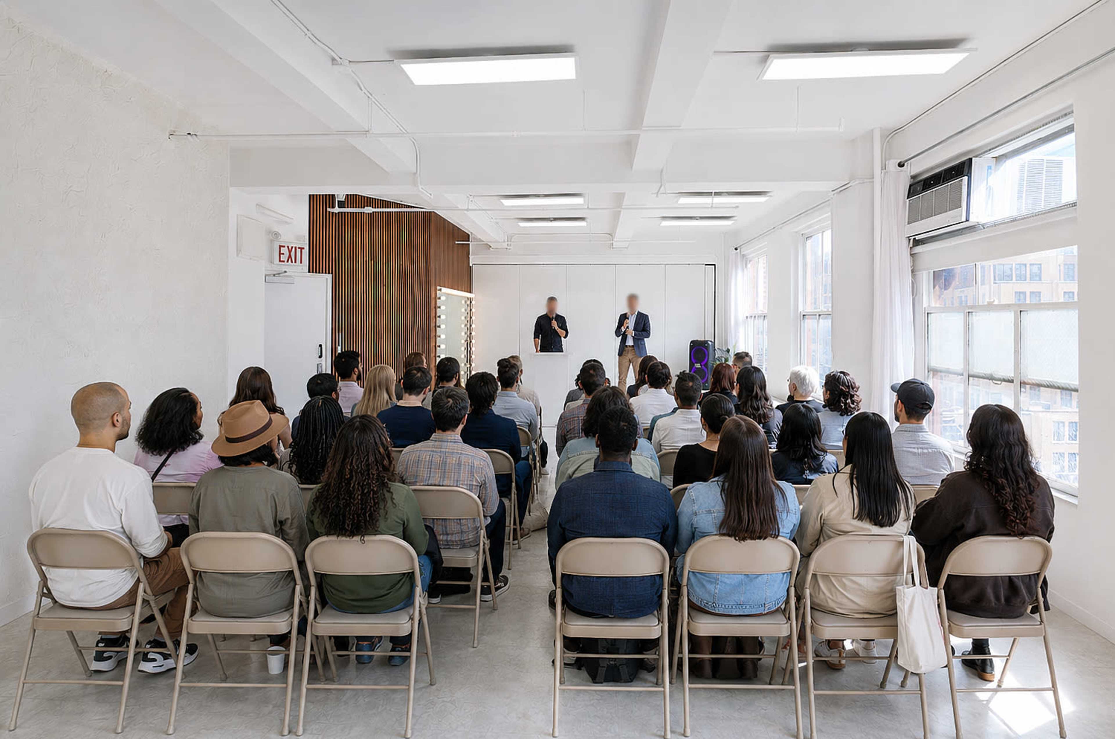 A diverse group of people is seated in a well-lit room, attentively watching two speakers in front of a presentation screen.