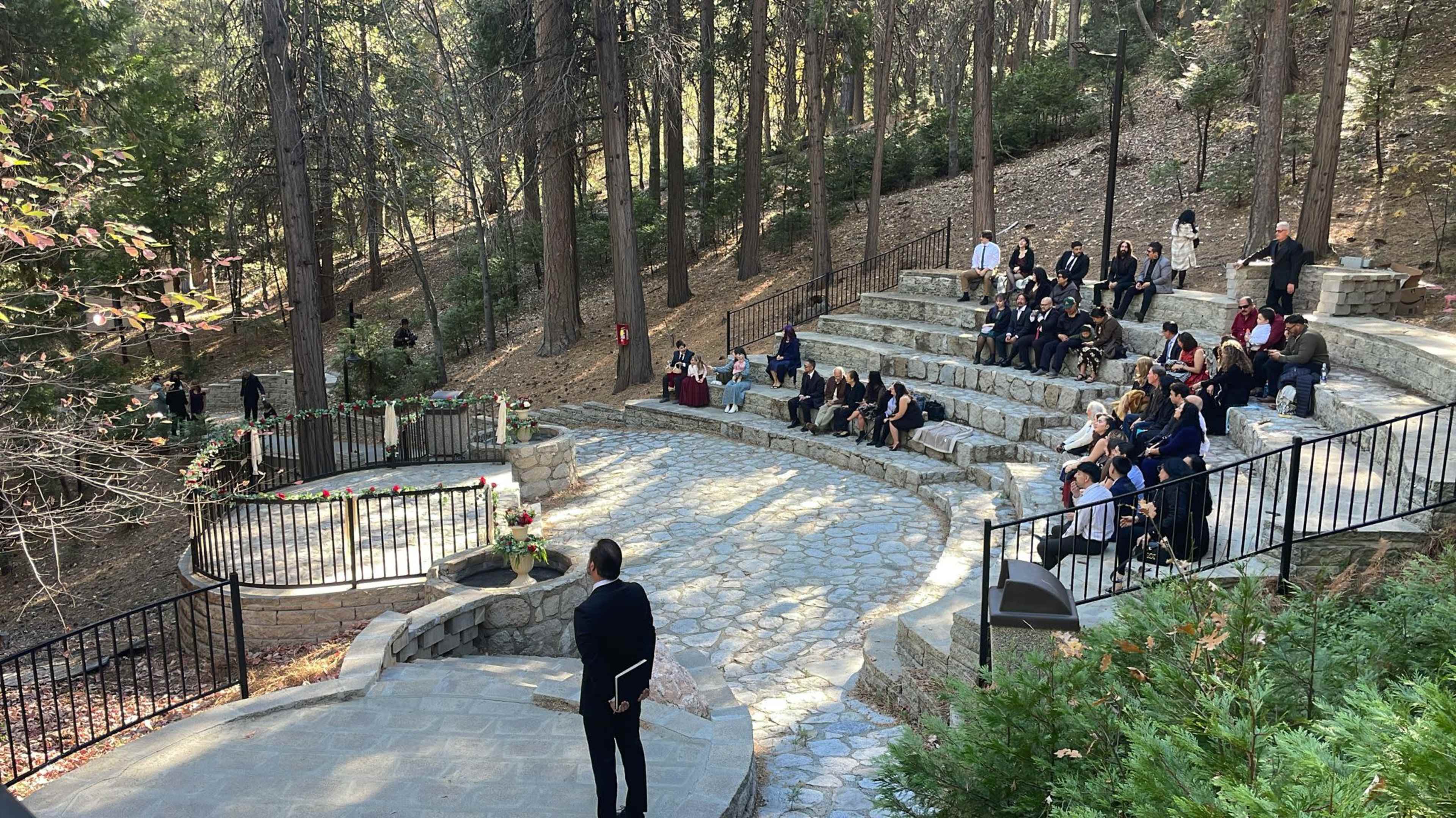 A formal outdoor gathering takes place in a stone amphitheater surrounded by trees, with attendees seated on the steps facing a speaker at the front.