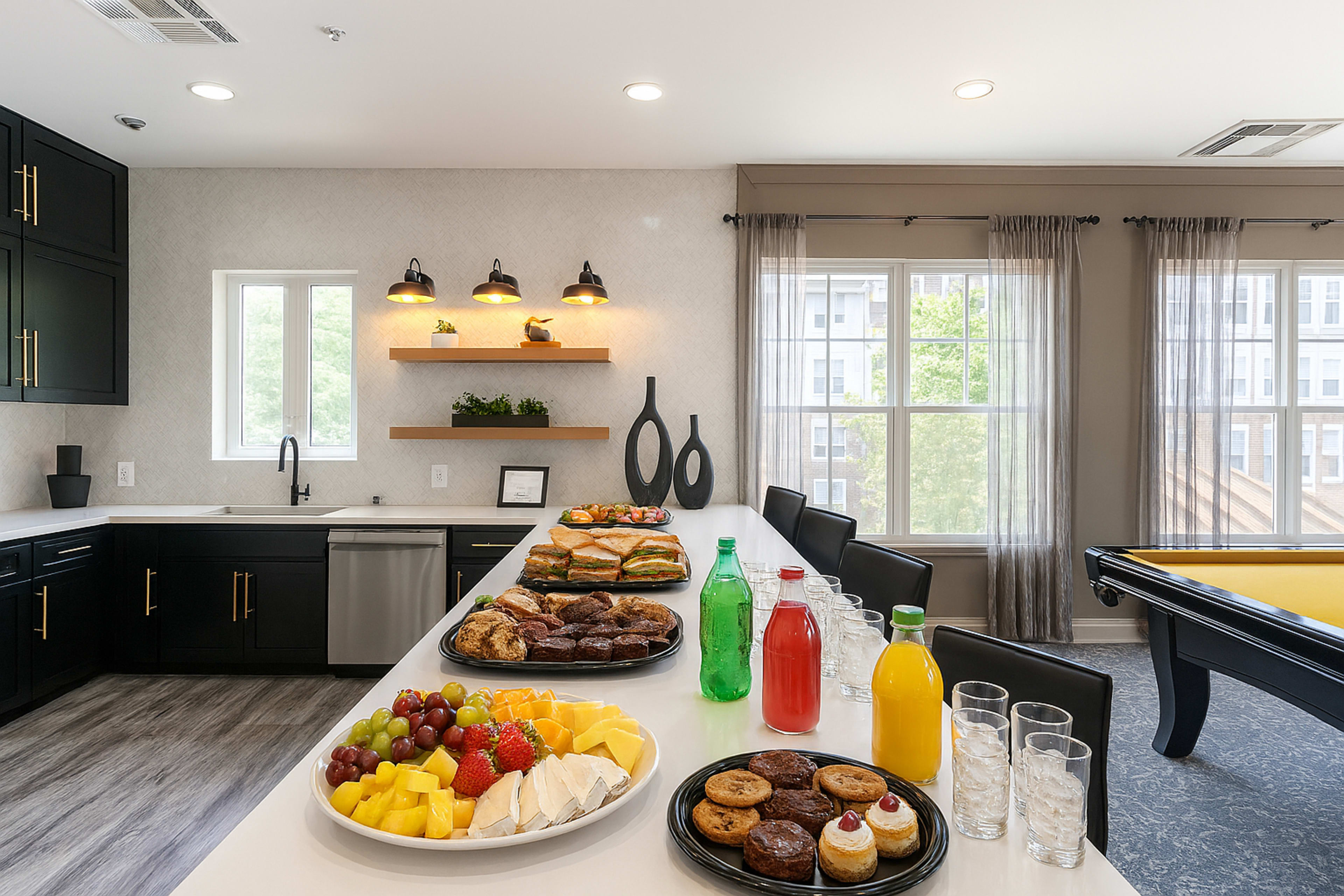 A buffet-style table displays a variety of foods, including fruits, pastries, and beverages, in a modern kitchen and dining area with large windows.