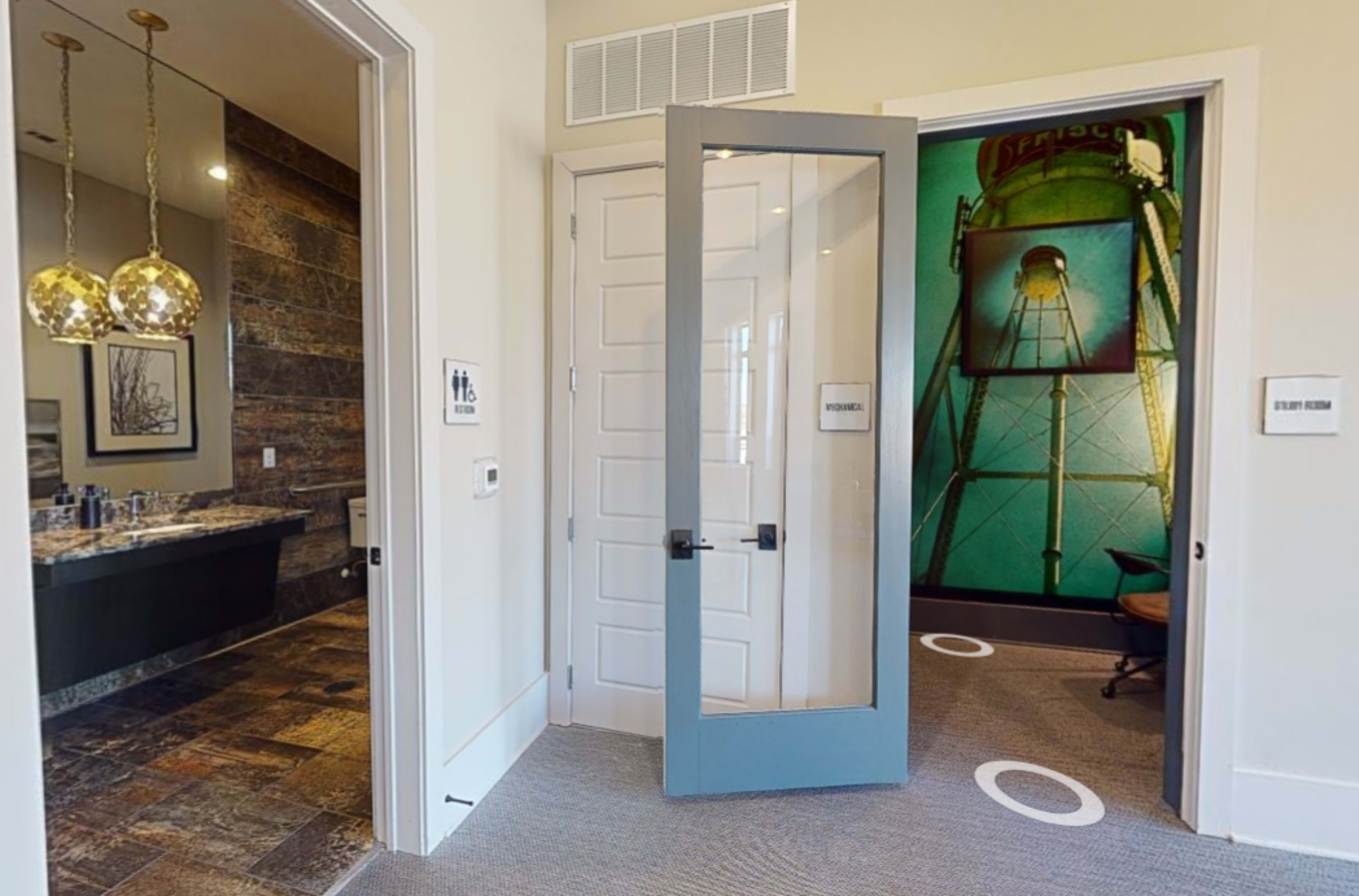 The image shows a hallway with a bathroom entrance on the left and a door leading to another room straight ahead, with a large wall art featuring a water tower.