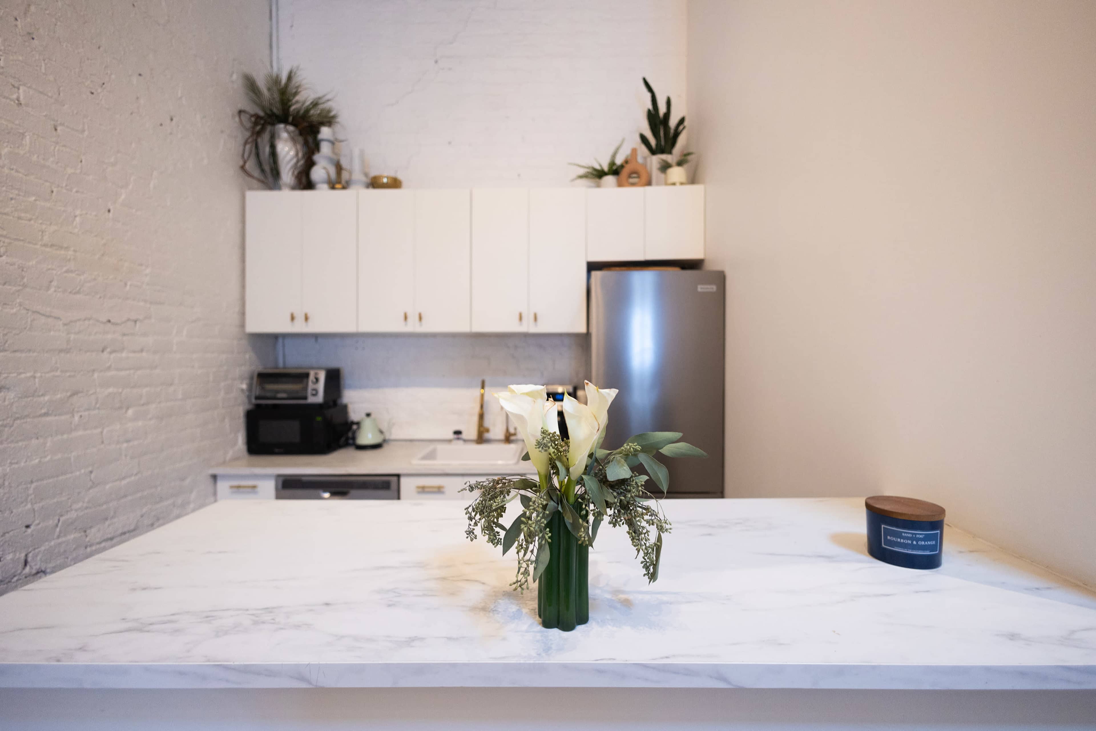 A kitchen features a white marble countertop with a vase of flowers, white cabinets, and a stainless steel refrigerator in the background.