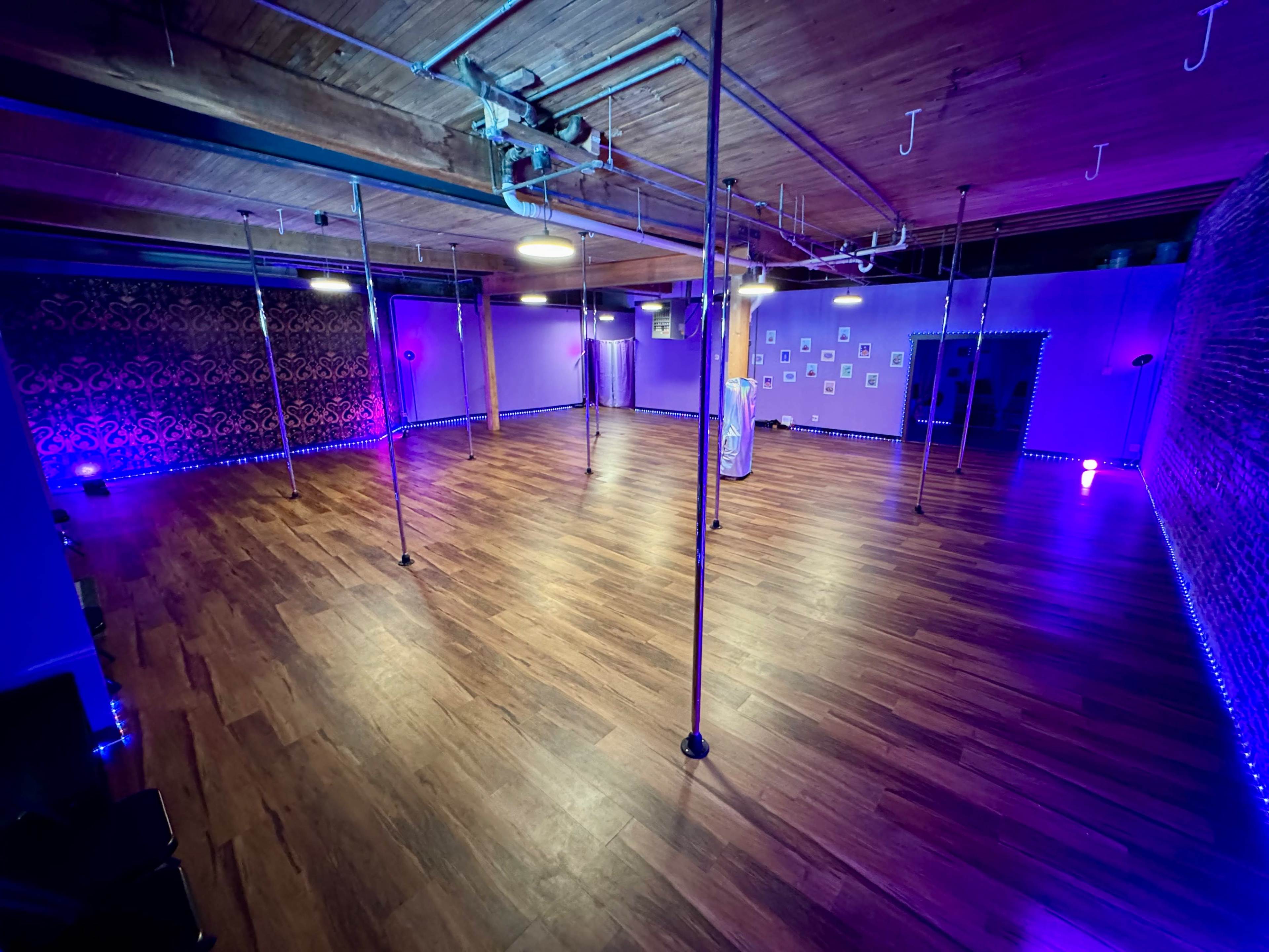 An empty dance studio with wooden flooring, multiple polished poles, and purple ambient lighting.