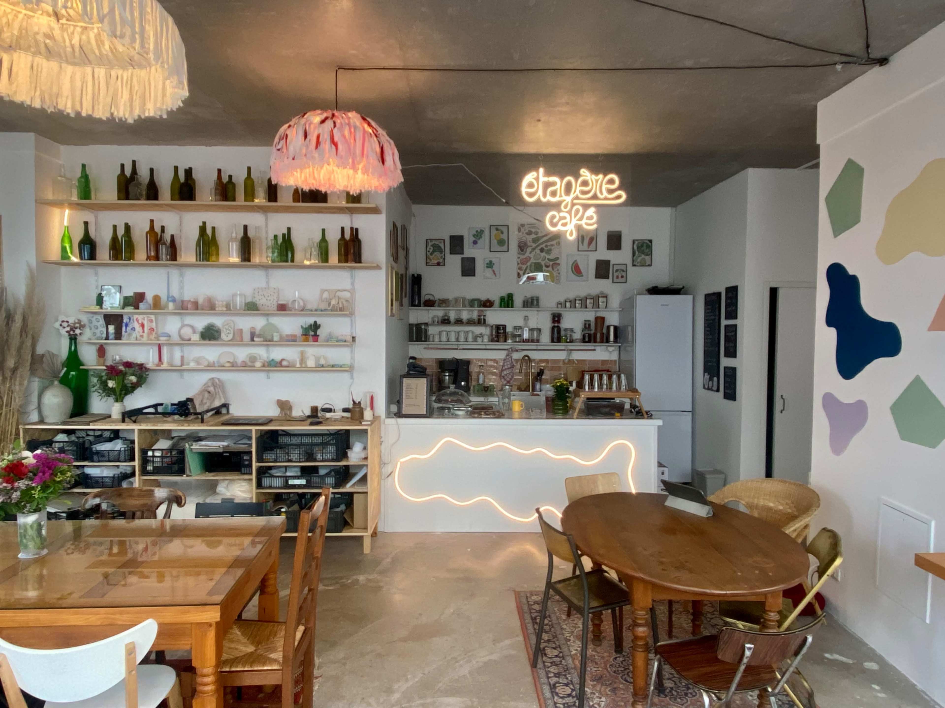 A cozy café interior with wooden tables, light fixtures, a counter featuring a neon sign, and various decorative items on shelves.