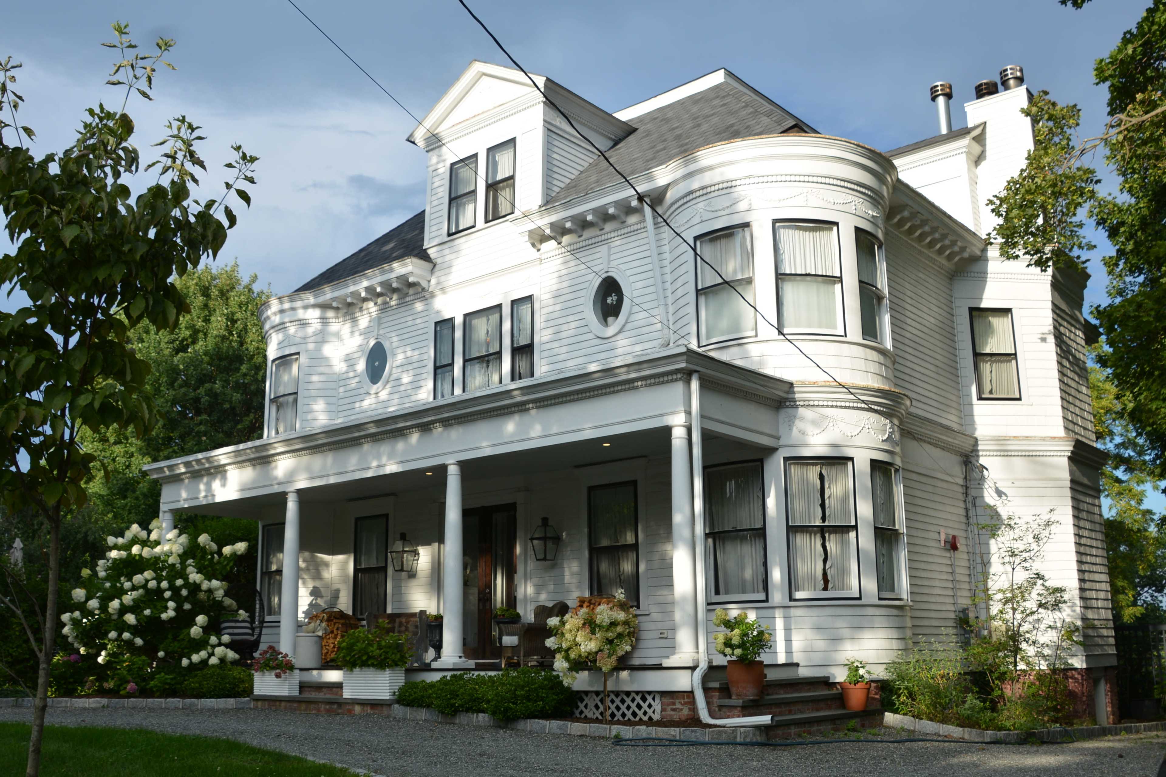 A large, two-story white house with a curved bay window and a front porch adorned with potted plants.