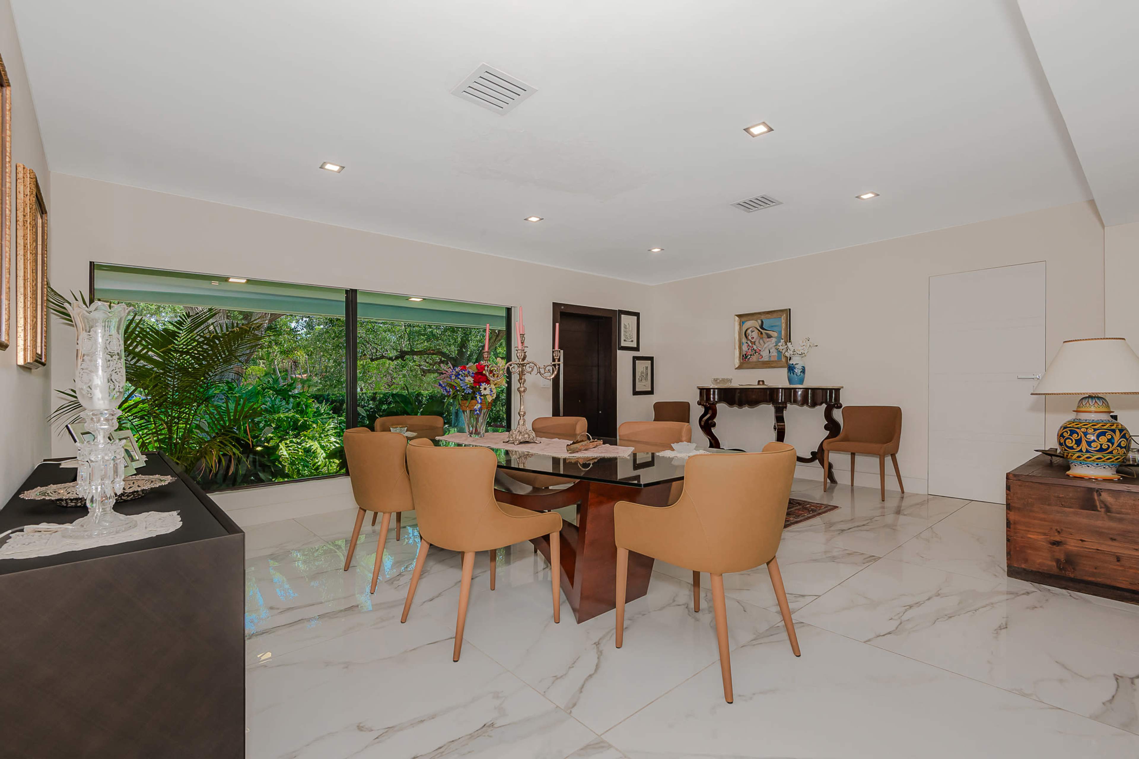 The image shows a modern dining area with a glass table surrounded by brown chairs, large windows offering views of greenery, and decorative elements on the walls and furniture.