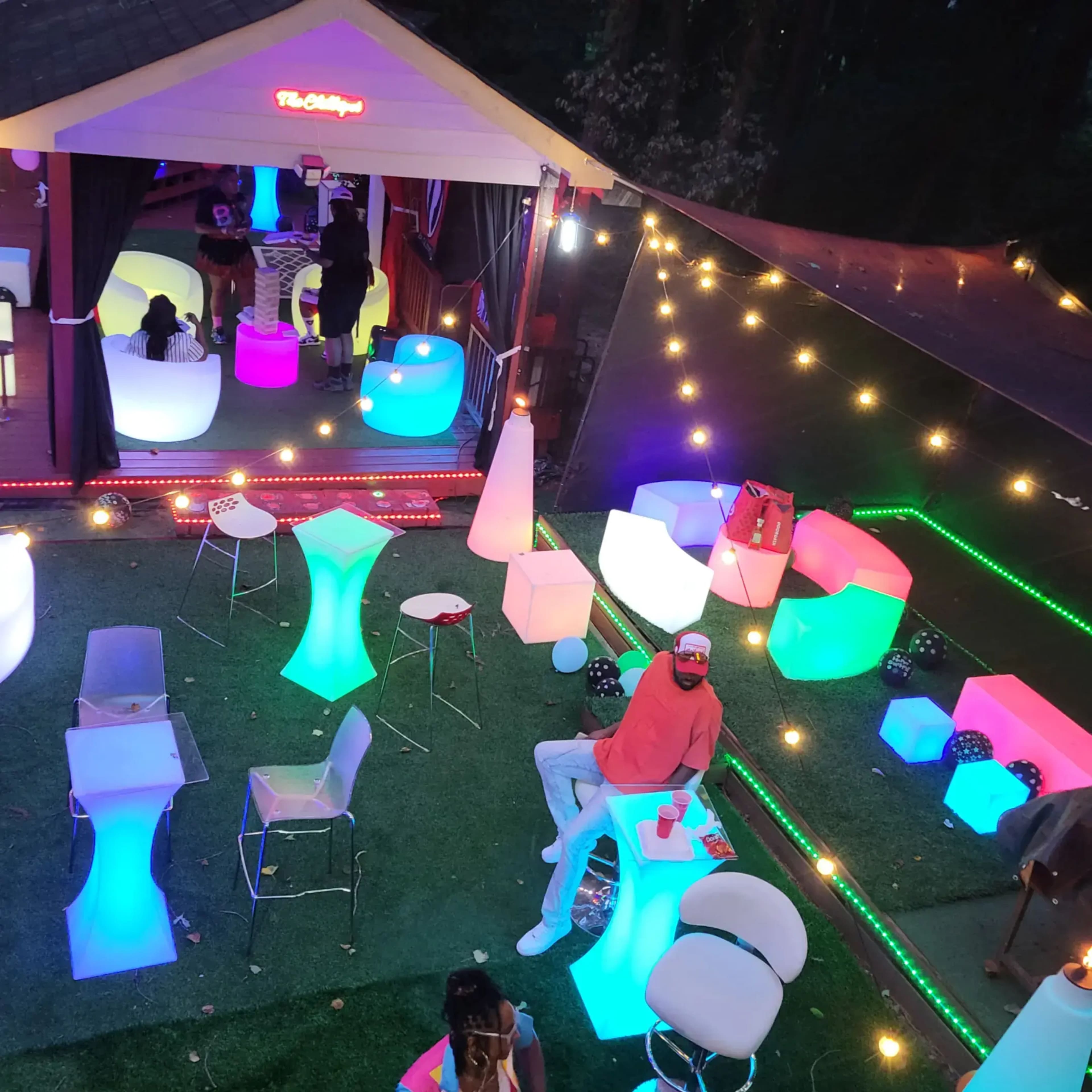 The scene shows a vibrant outdoor lounge area illuminated by colorful LED furniture and string lights, with people mingling and seated at various glowing tables and chairs.