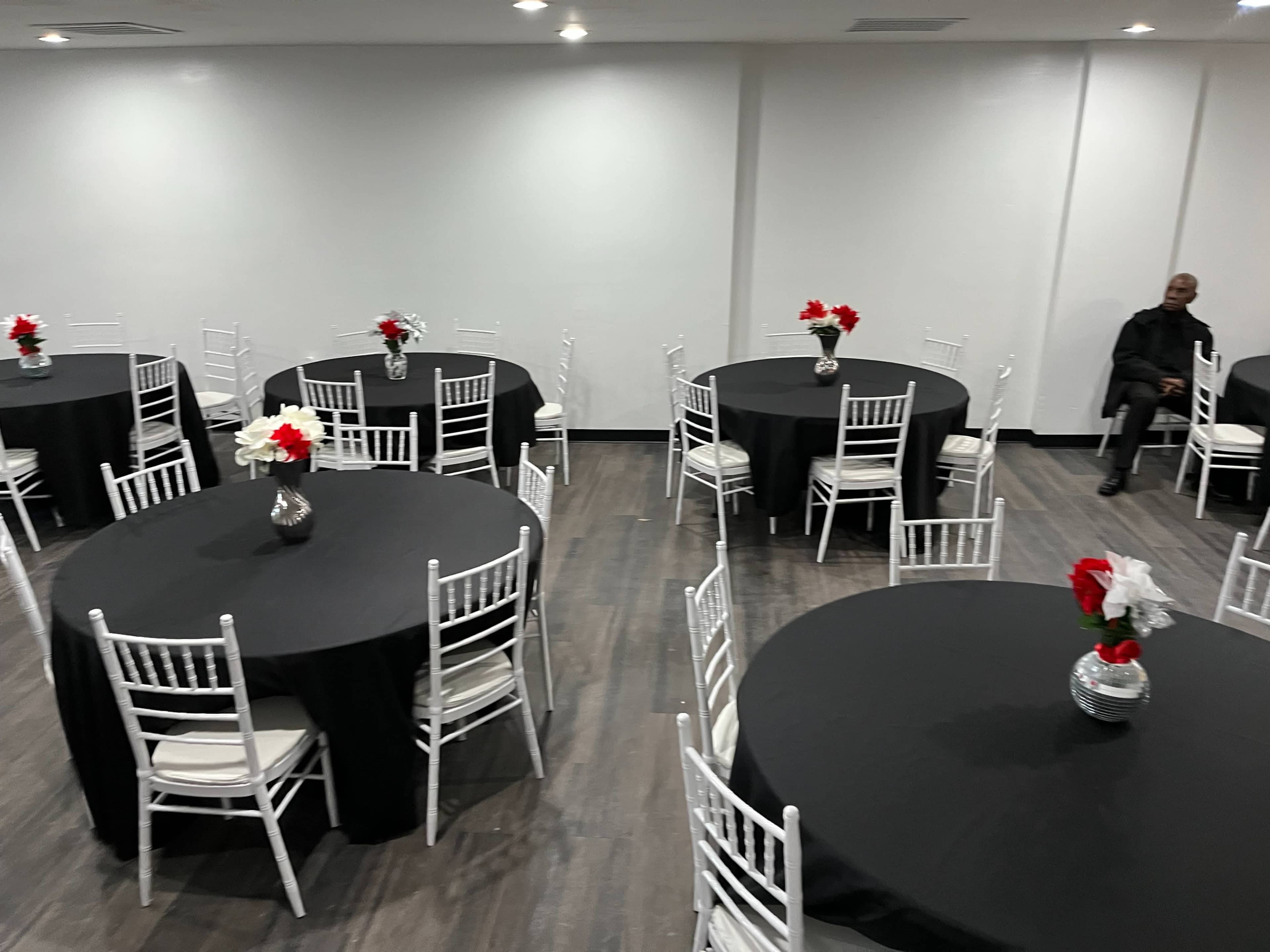 A large event space features several round tables covered with black tablecloths, each adorned with floral centerpieces, while a lone individual sits at the edge of the room.