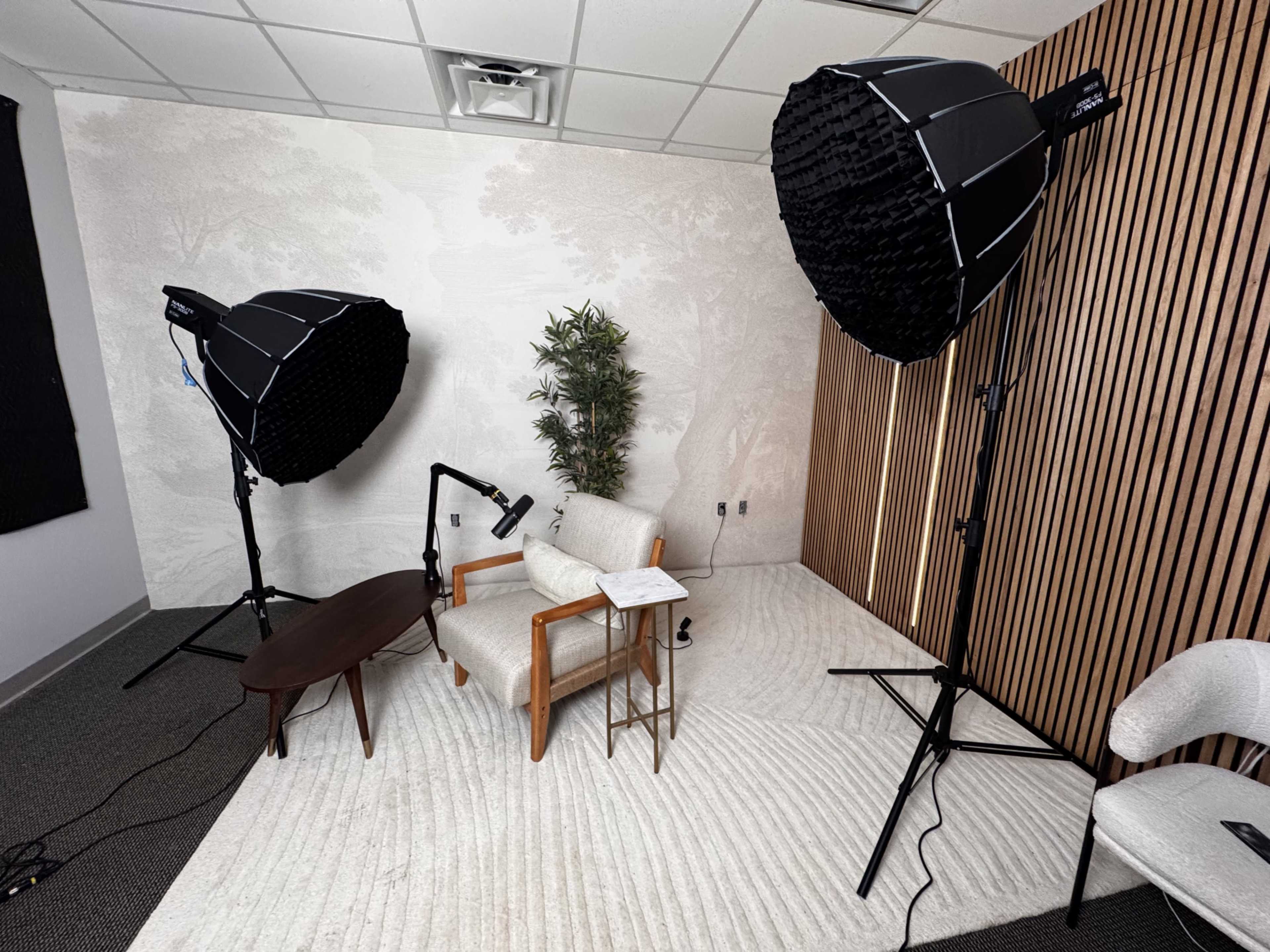 The image shows a studio setup with two large softbox lights, a wooden table, an armchair, and a plant against a textured wall.