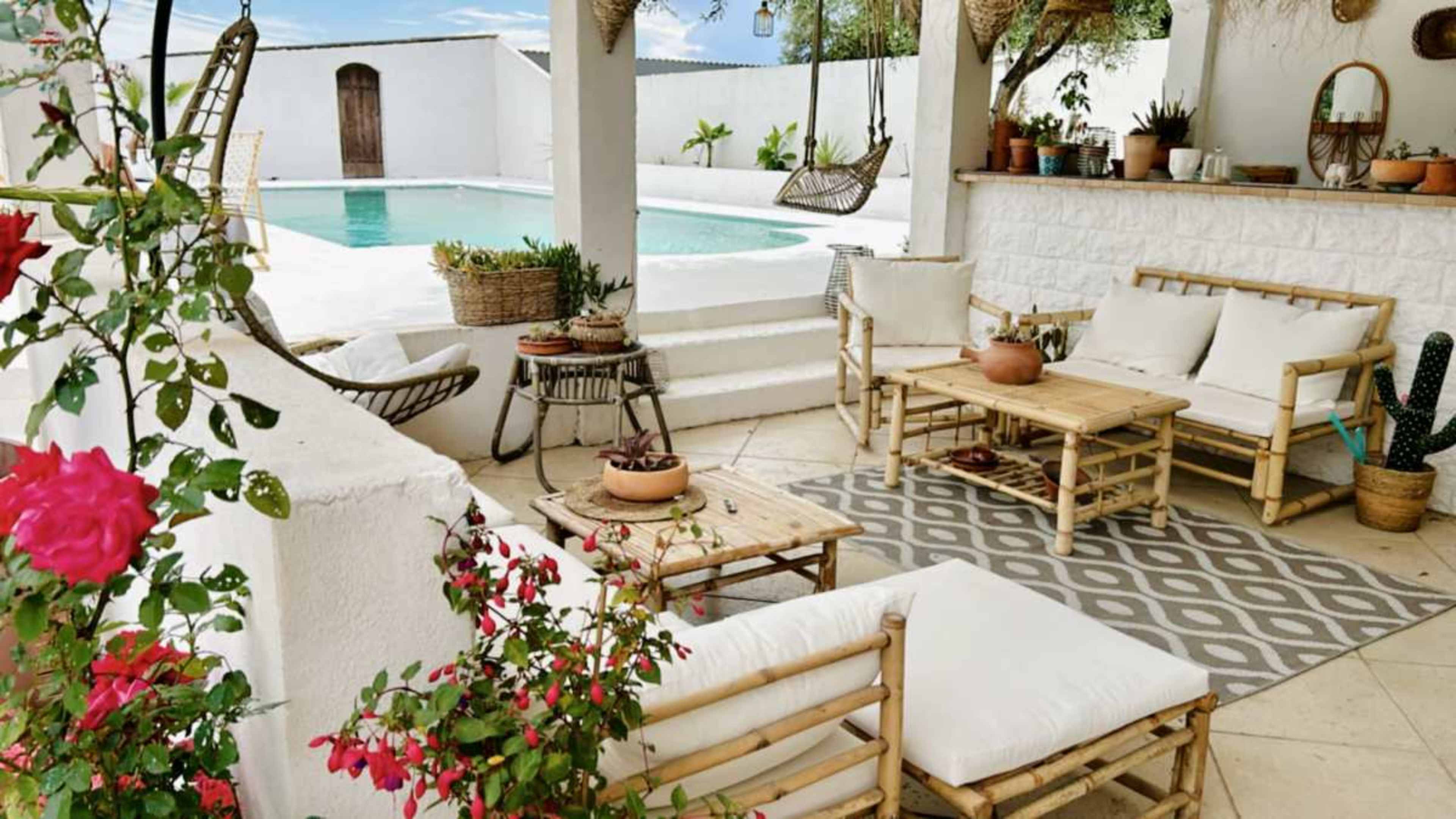 The image shows a bright outdoor patio with bamboo furniture, a patterned rug, potted plants, and a swimming pool in the background.