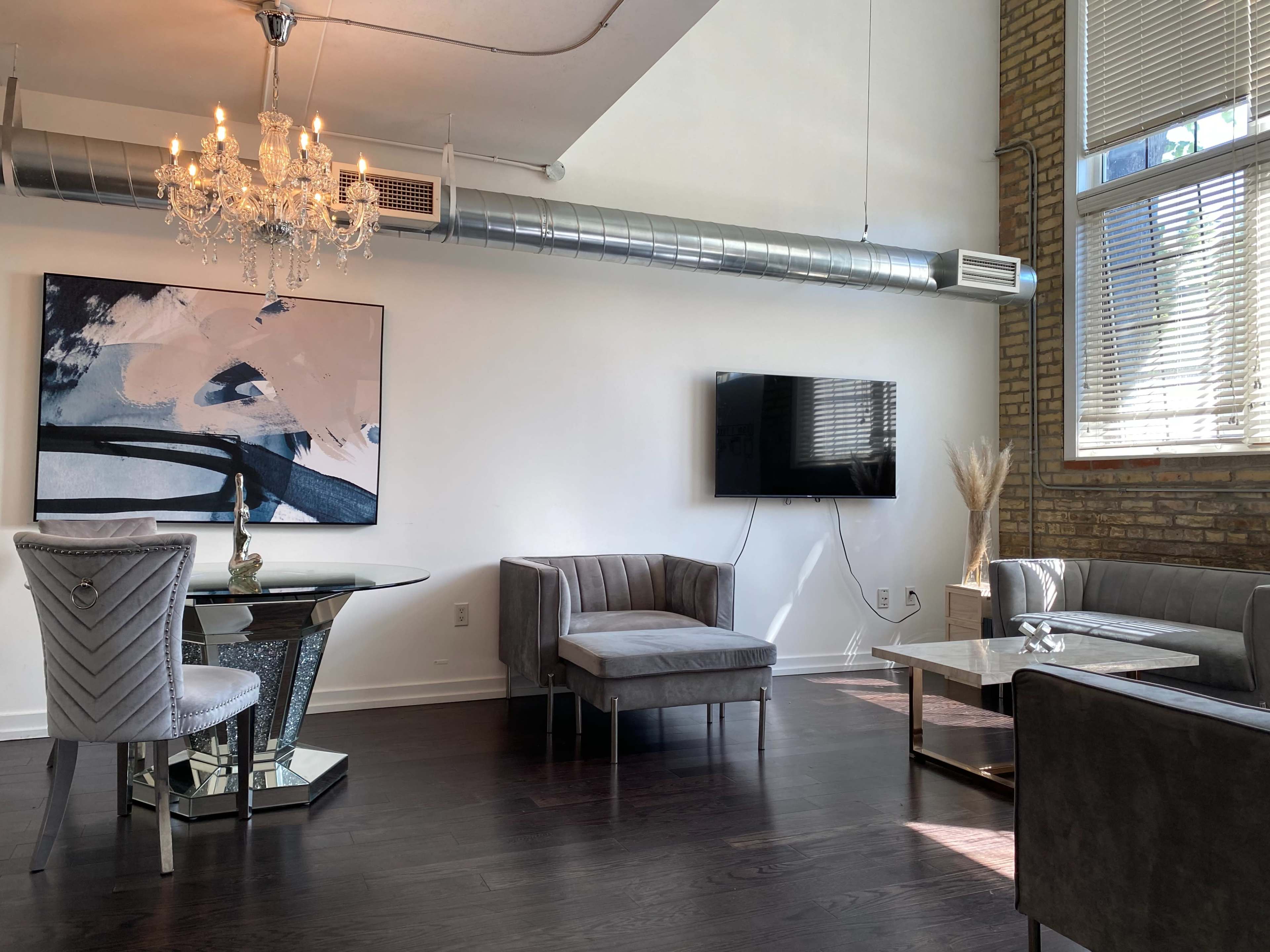 The image shows a modern living space featuring a glass dining table, a velvet chair, a sleek sofa, and a mounted television against a backdrop of exposed brick walls.