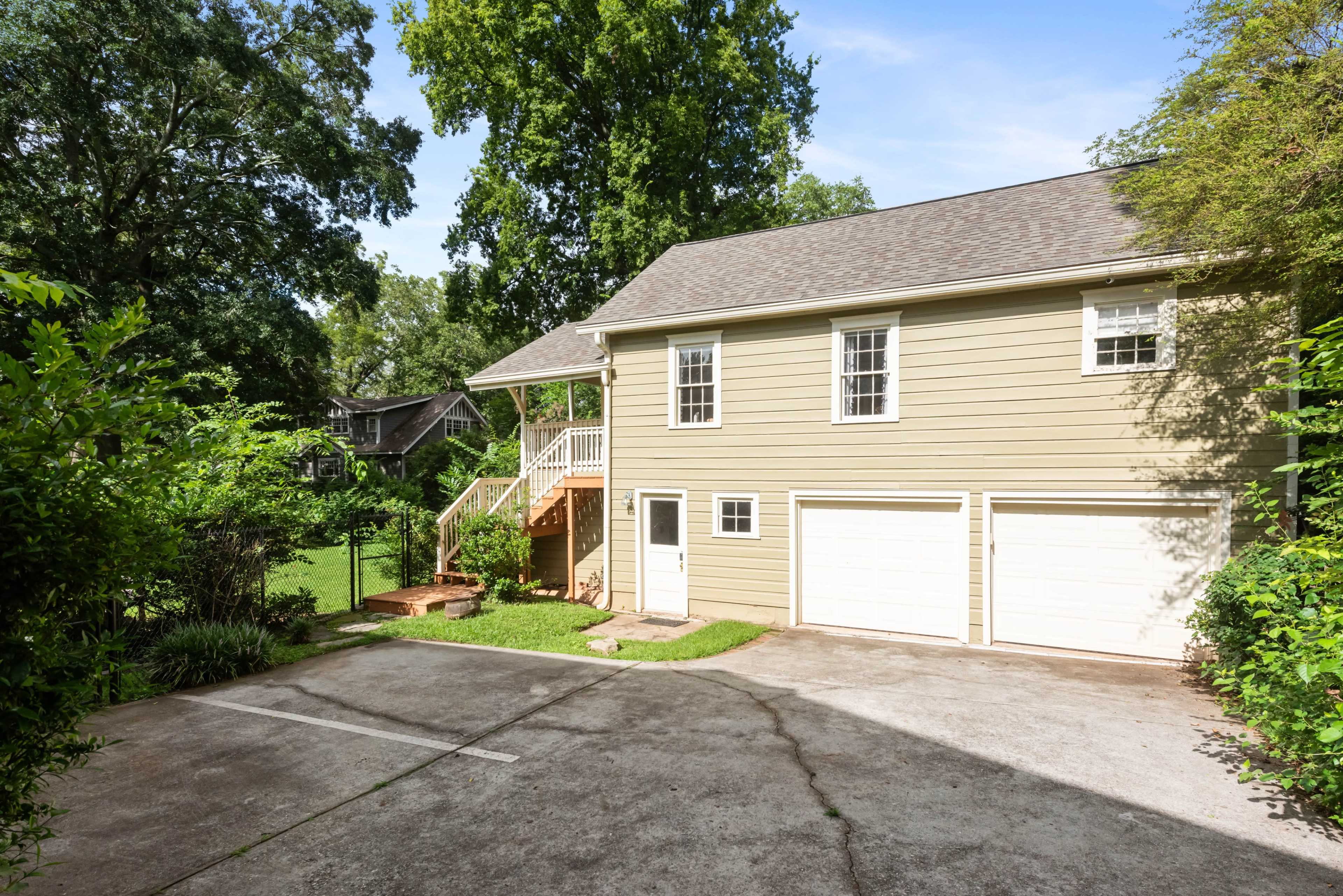The image shows a two-story house with a set of stairs leading to a porch, situated next to a driveway and surrounded by greenery.