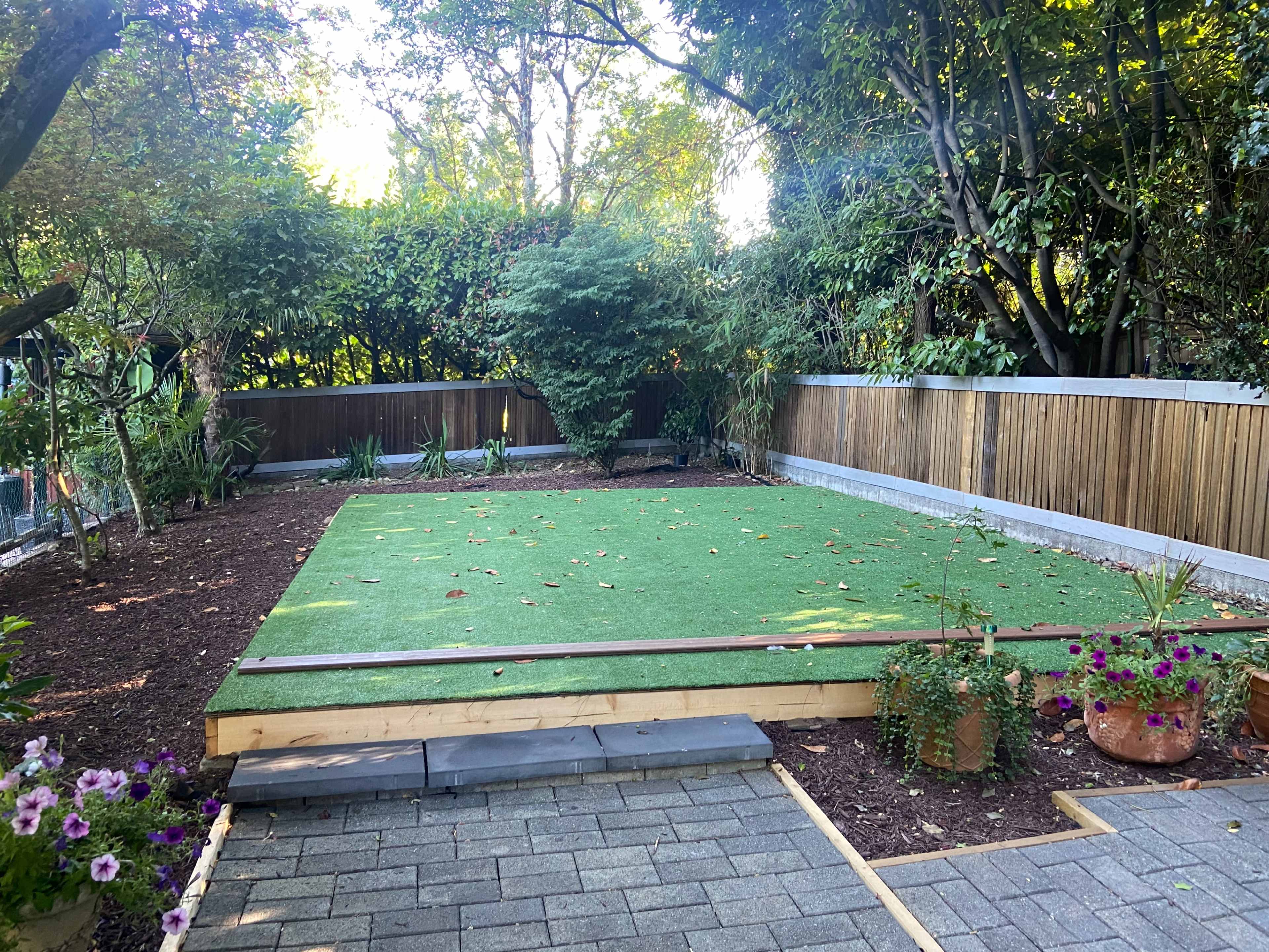A landscaped backyard featuring a rectangular area of artificial turf surrounded by wooden fencing, plants, and potted flowers.