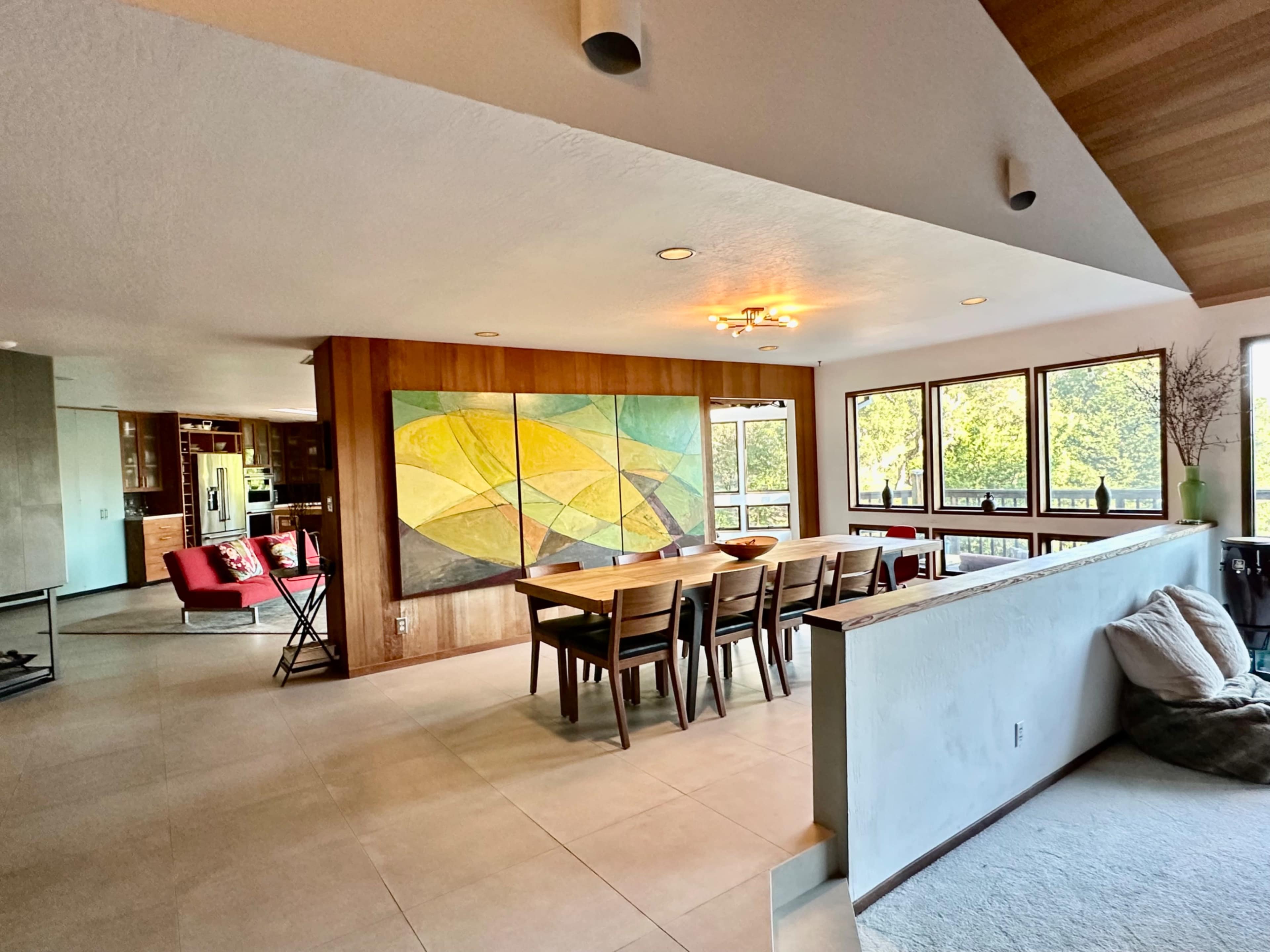 The image shows a modern interior space featuring a dining area with a large abstract painting, surrounded by large windows that provide natural light.