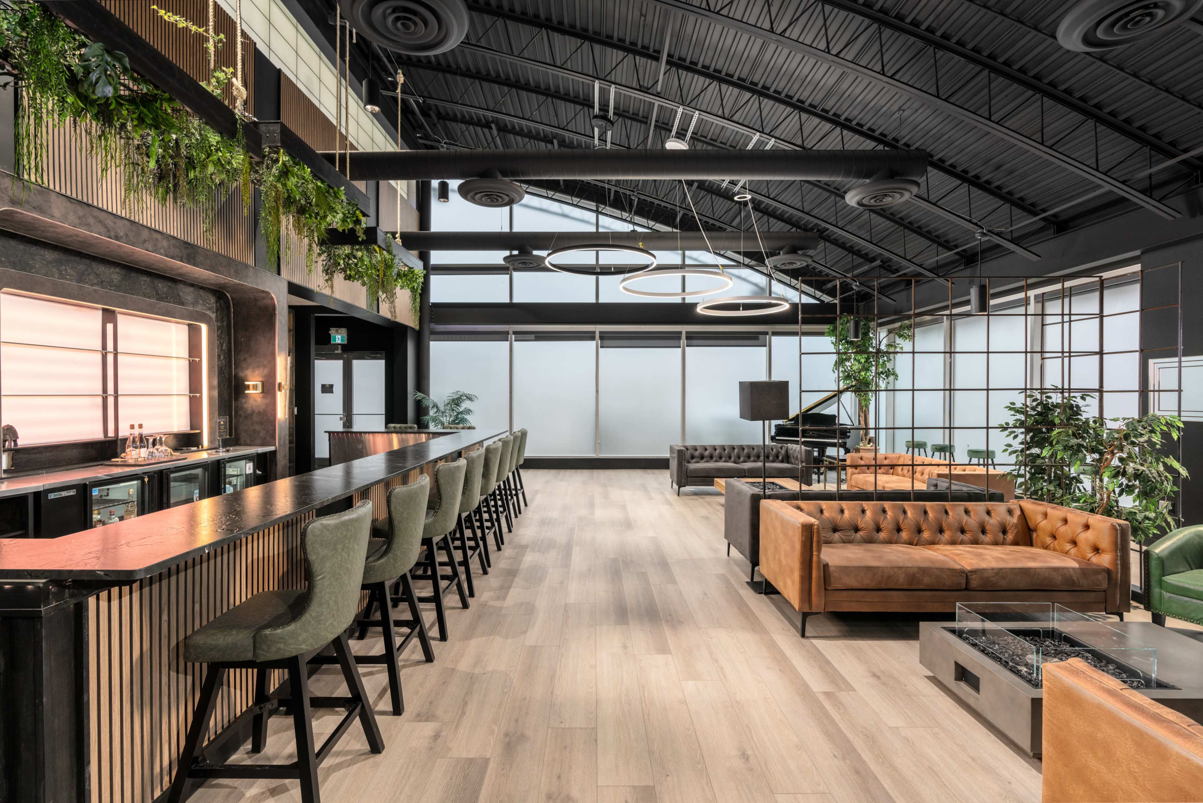 A modern interior space featuring a bar with seating, a lounge area with leather sofas, and various plants throughout the design.