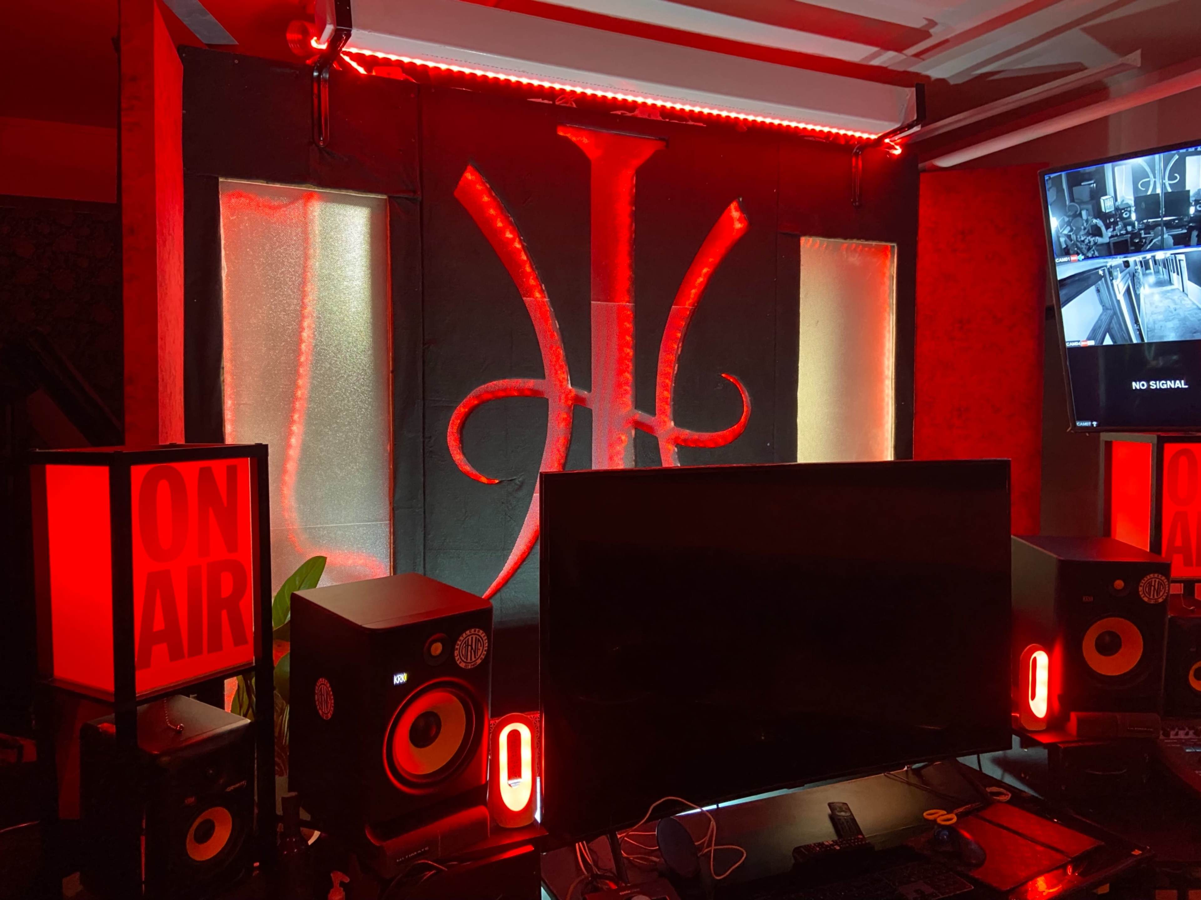 The image shows a music or broadcast studio with a glowing red backdrop featuring a symbol, accompanied by speakers, a monitor, and decorative lighting.