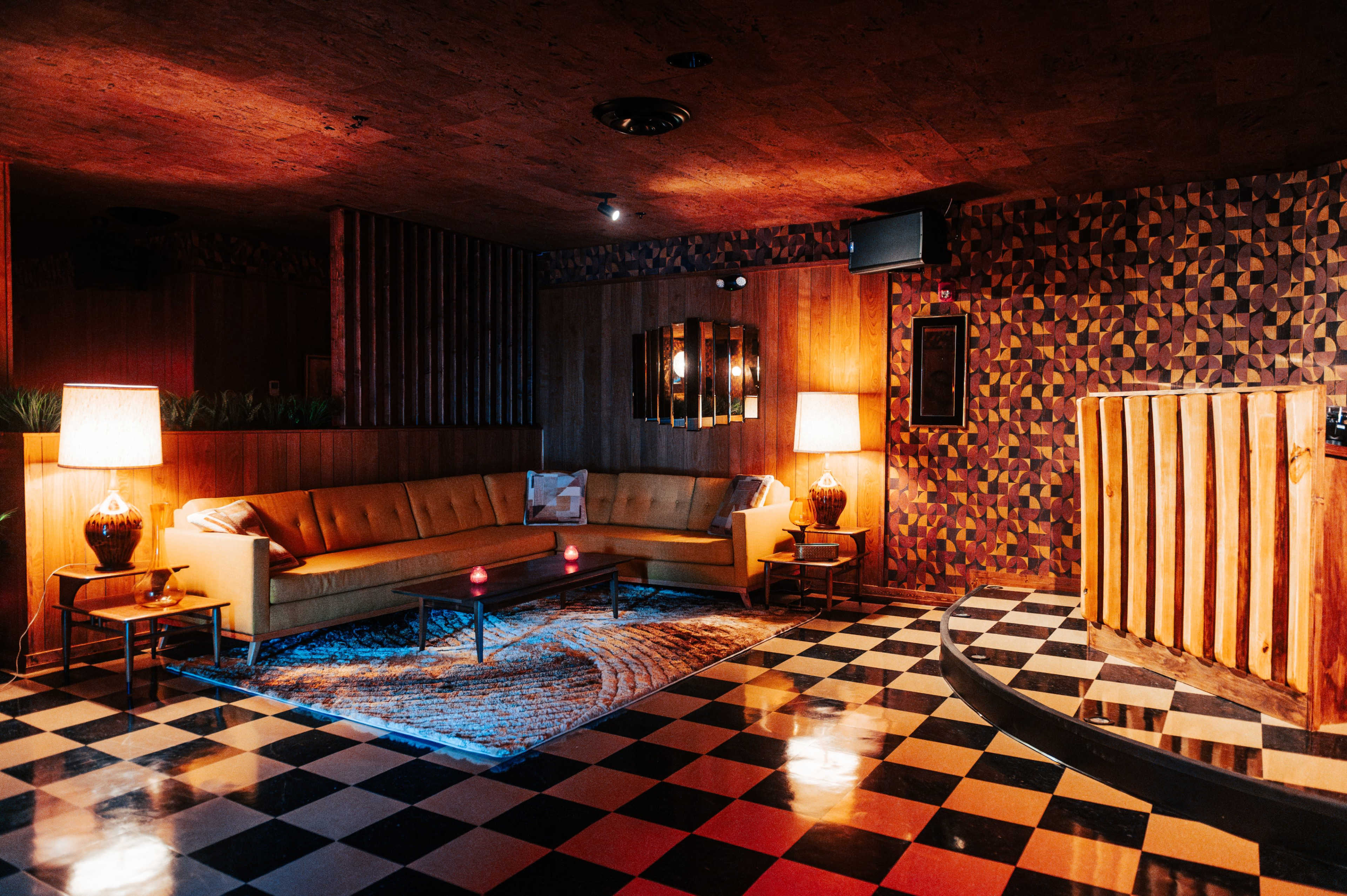 The image shows a retro-style lounge with a checkered floor, a large yellow sofa, and two lamps illuminating the space.
