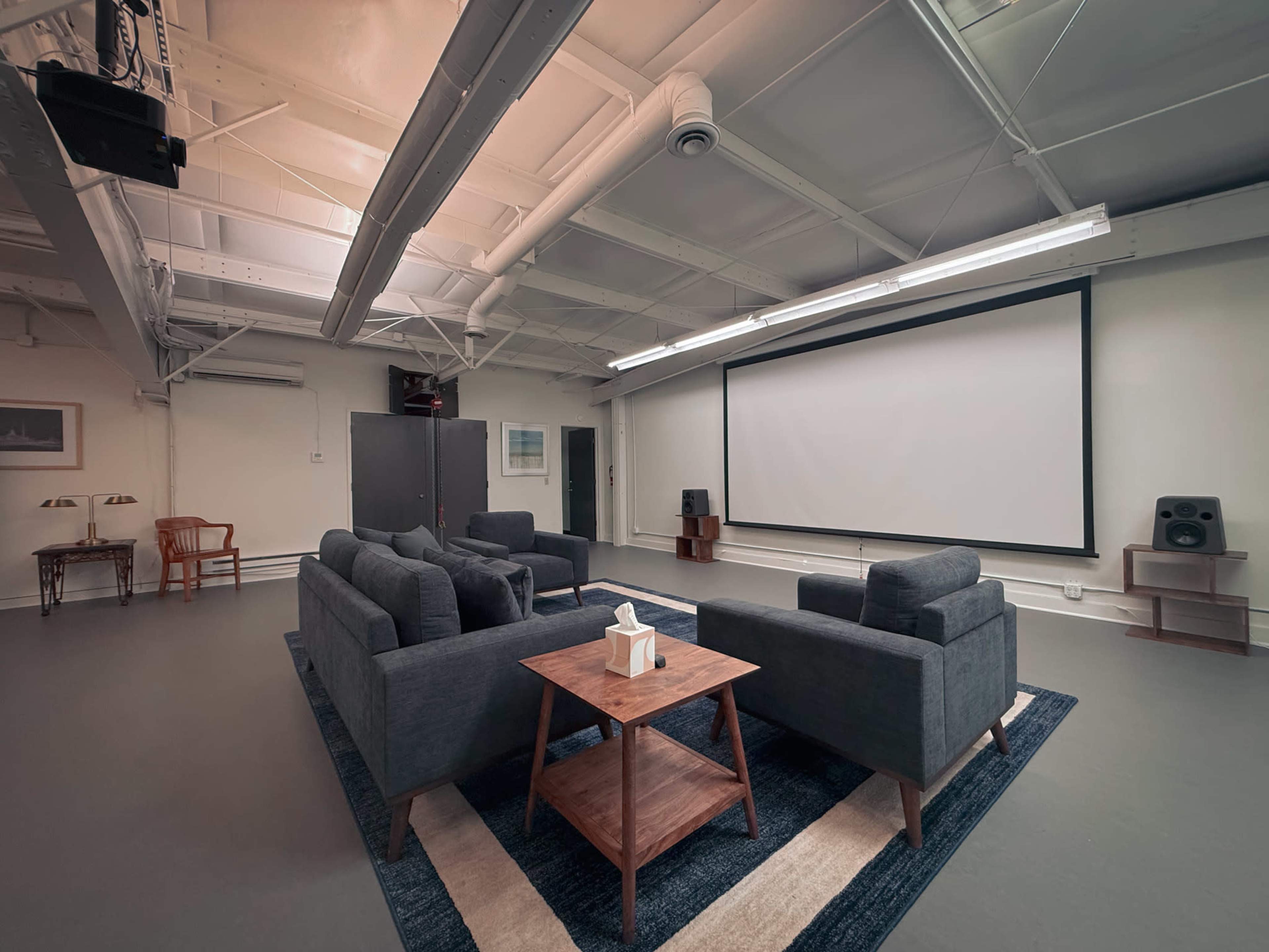 A spacious, well-lit room features a large projection screen, two gray sofas, a wooden coffee table, and audio equipment, with a minimalist design.