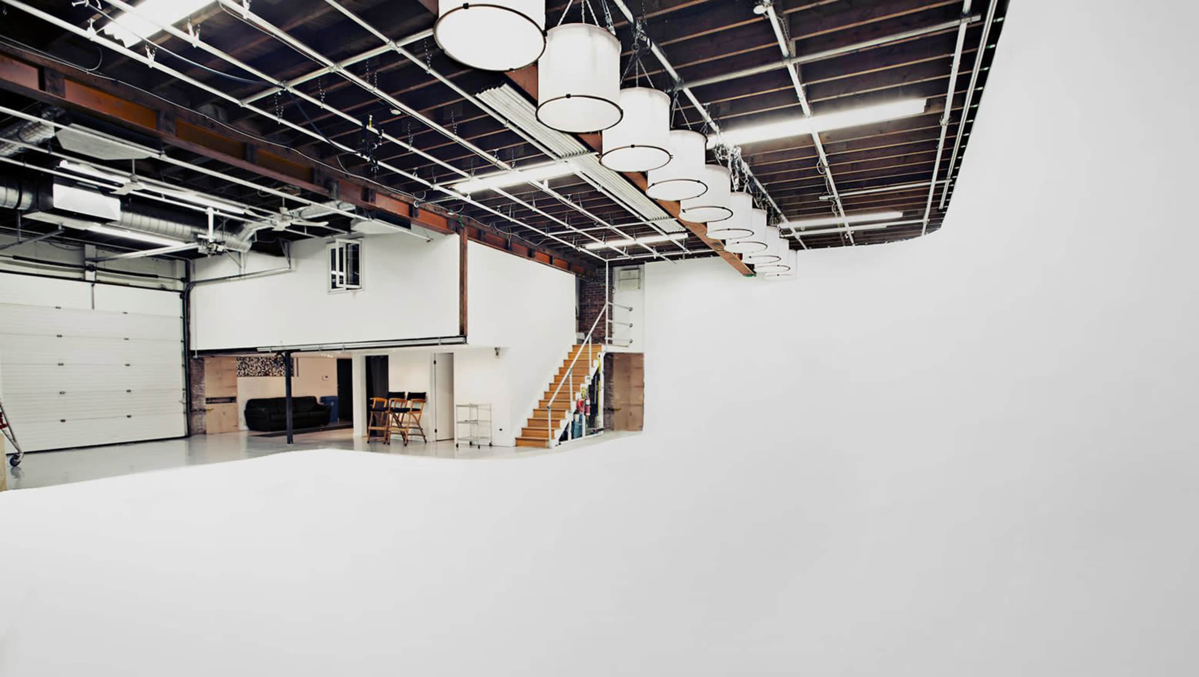 The image shows a spacious studio with a white cyclorama wall, overhead lighting, and a staircase leading to an upper level.