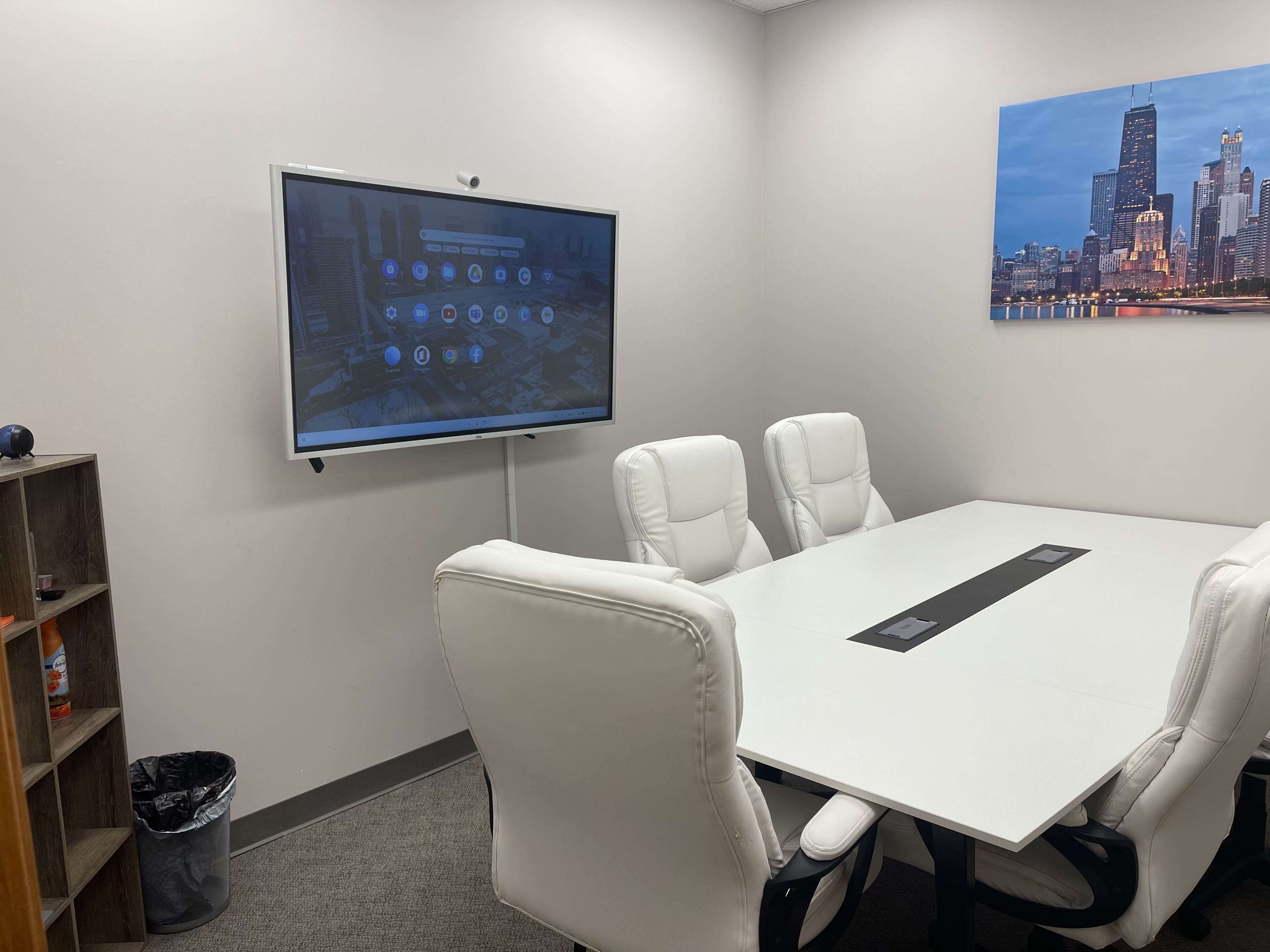 A modern conference room with a large screen displaying a digital interface, six white chairs around a rectangular table, and a small shelf with items in the corner.