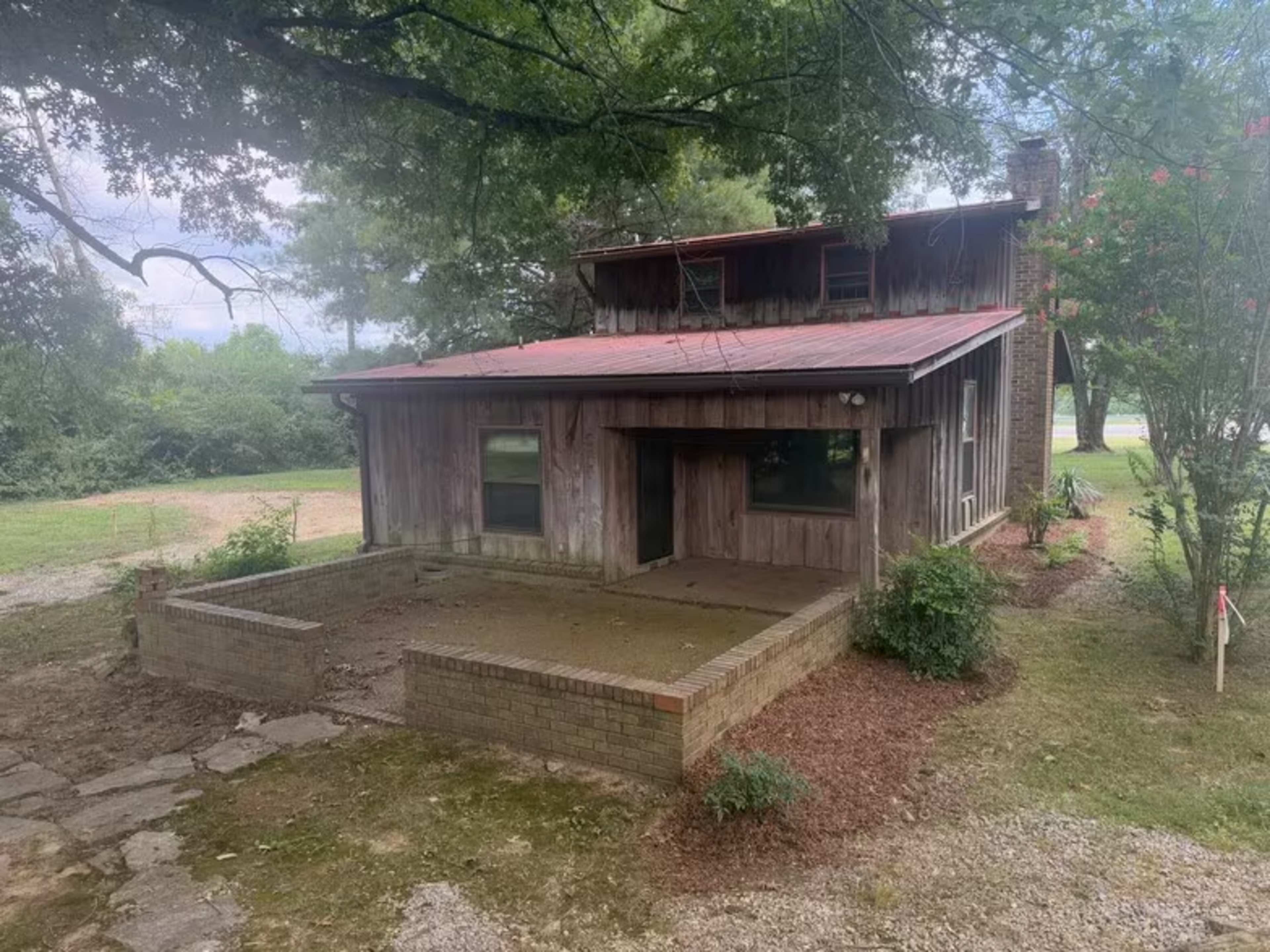 The image shows a weathered wooden house with a red metal roof and a small, enclosed patio area surrounded by landscaping.