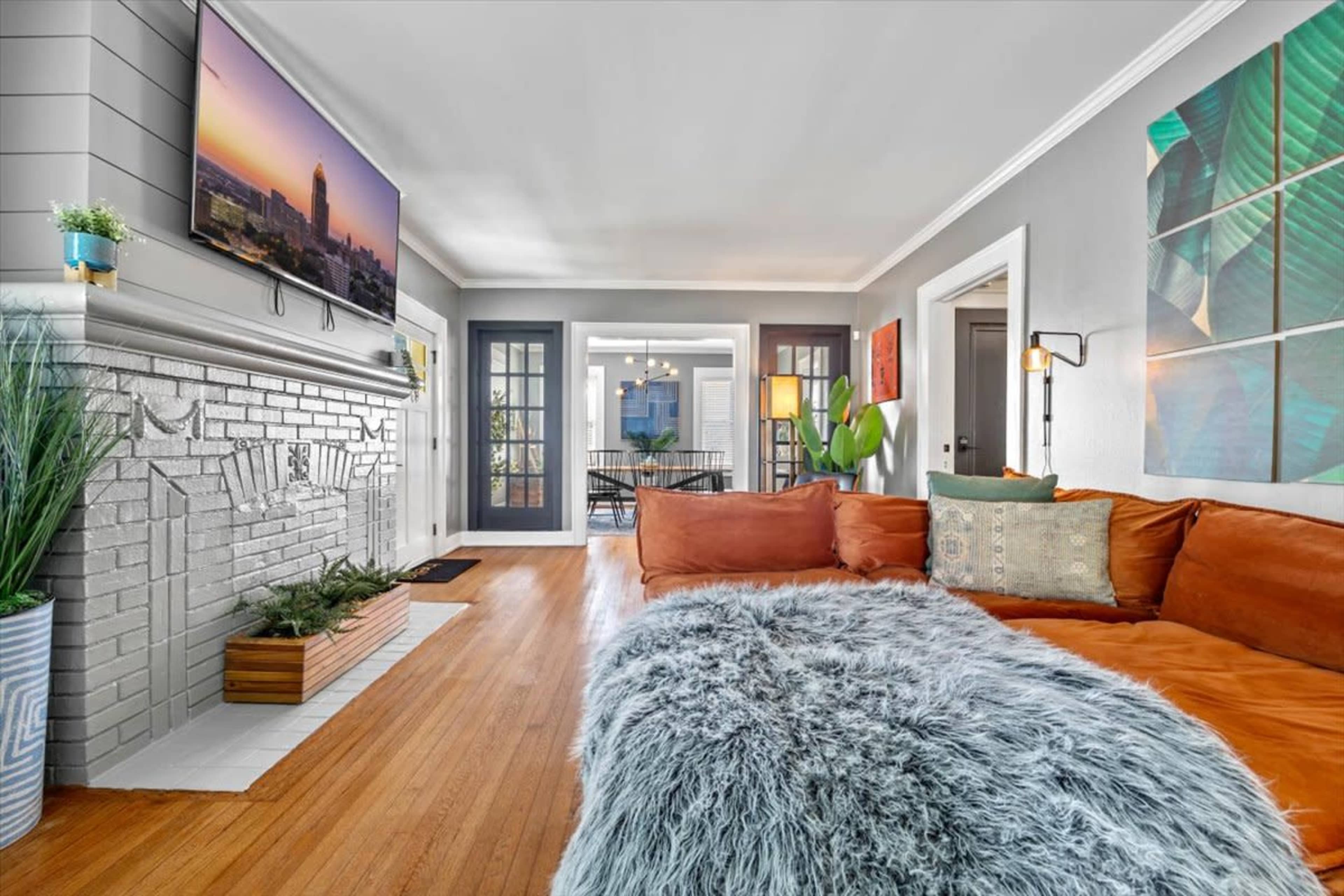 A cozy living room features a large orange sofa with a fluffy gray blanket, a wall-mounted TV, and doors leading to a dining area.