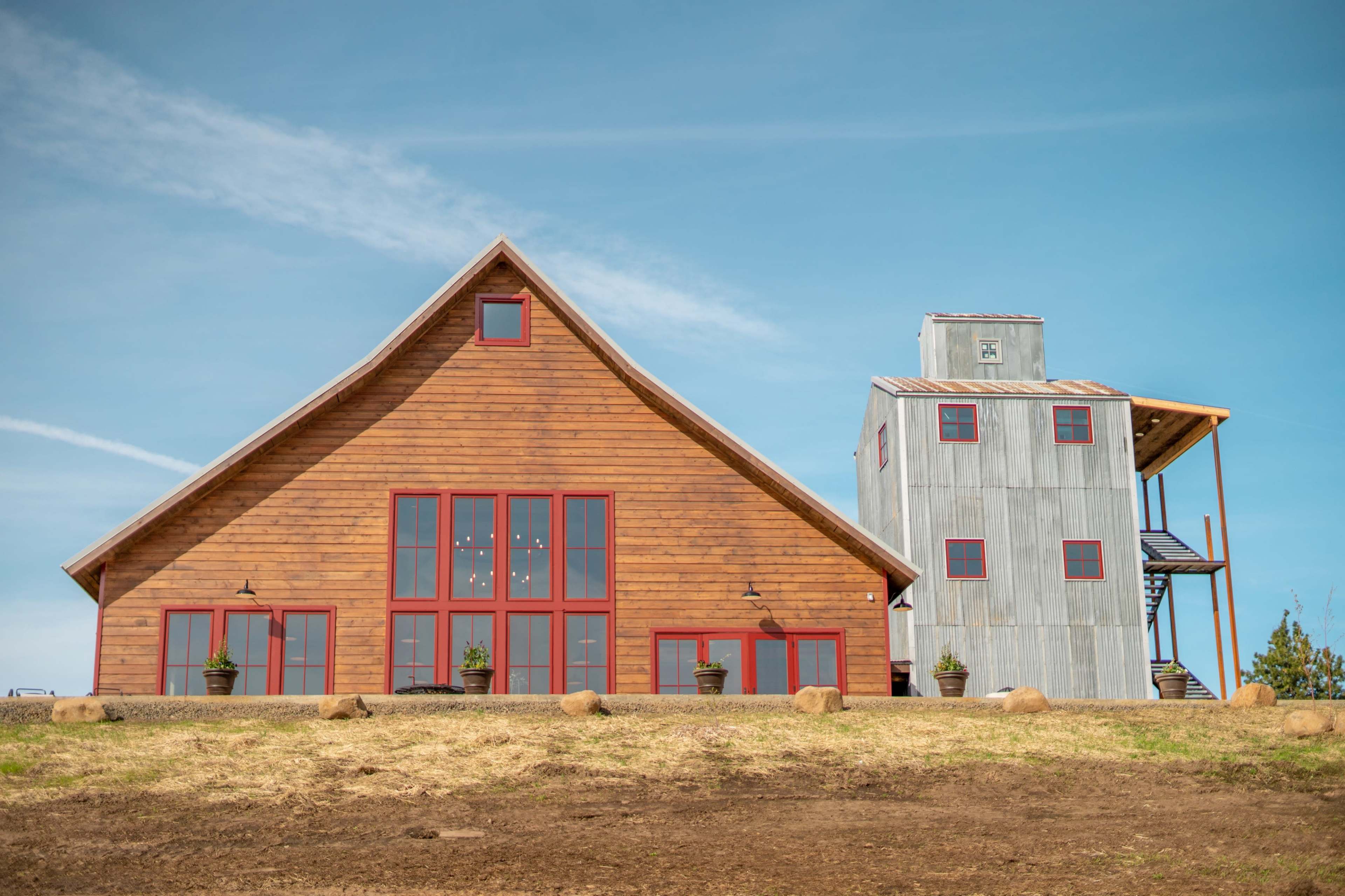 A large wooden barn-style building with red accents stands next to a gray metal structure on a grassy hillside.