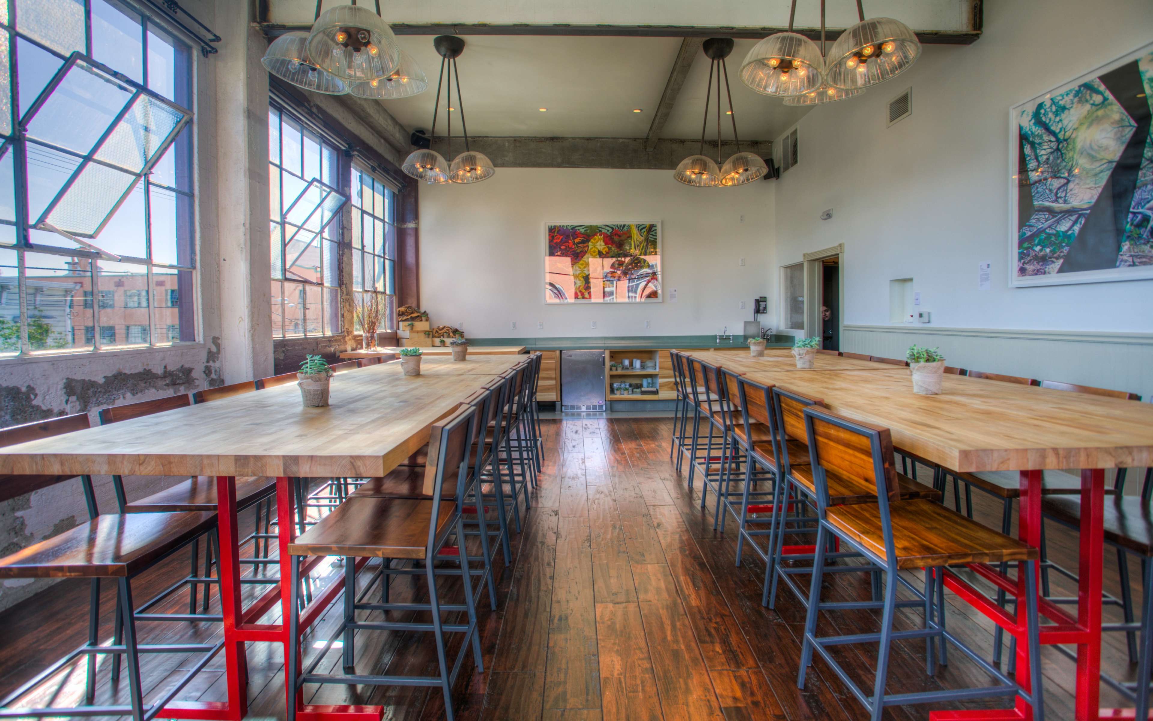 The image shows a modern dining area with long wooden tables, metal bar stools, large windows, and pendant lighting.
